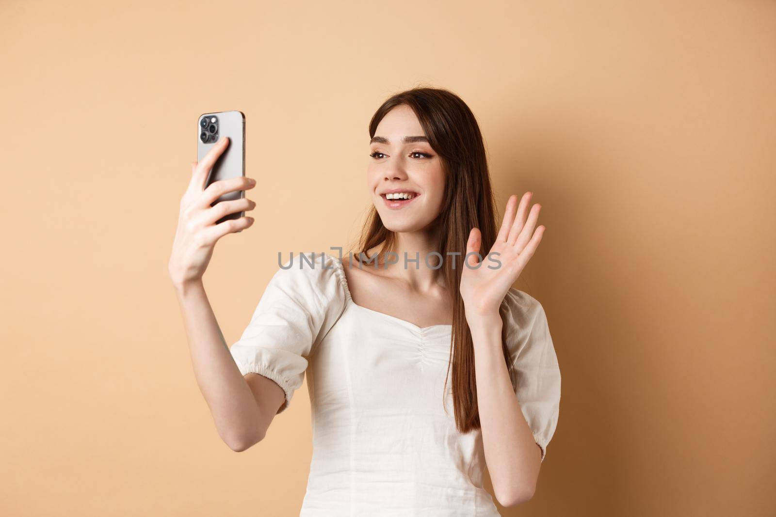 Girl video chat on phone, waiving hand at mobile camera and saying hello, talking to friend, smiling and standing on beige background.