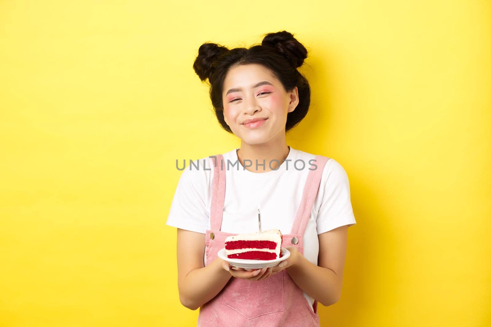 Happy asian birthday girl with bright makeup, blowing candle on cake, making wish, standing on yellow background by Benzoix