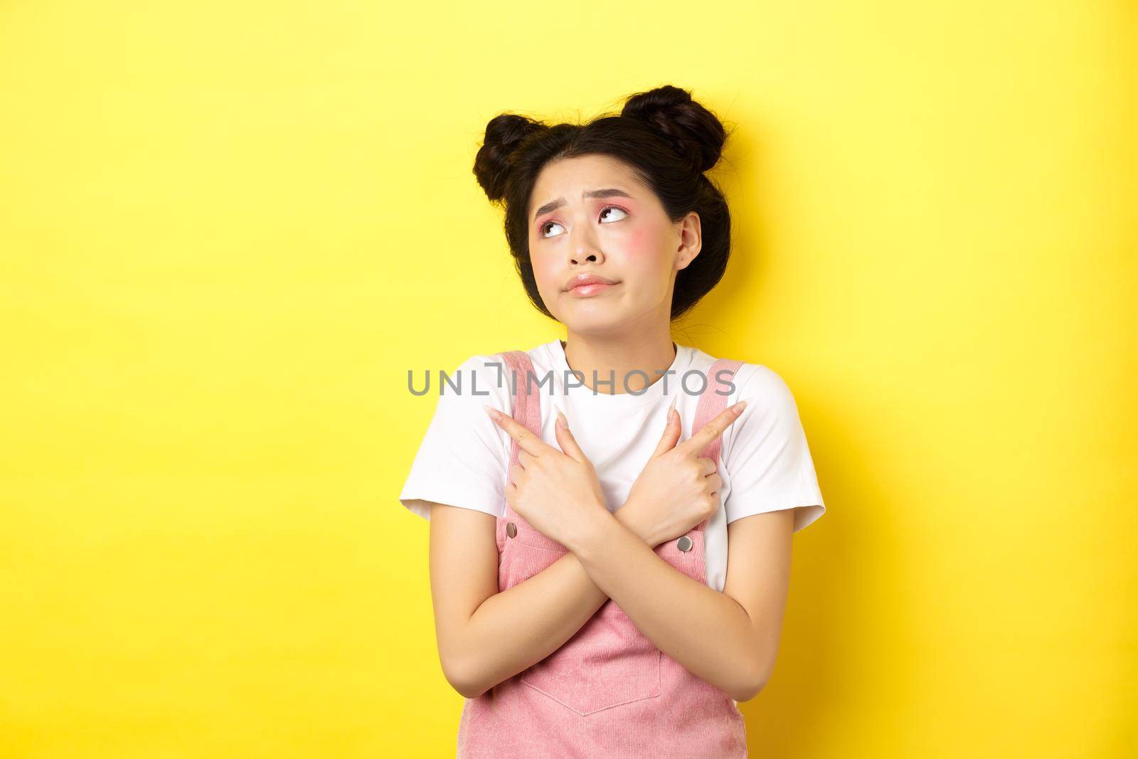 Indecisive asian teen girl troubled to make choice, pointing sideways and looking at logo confused, standing on yellow background.