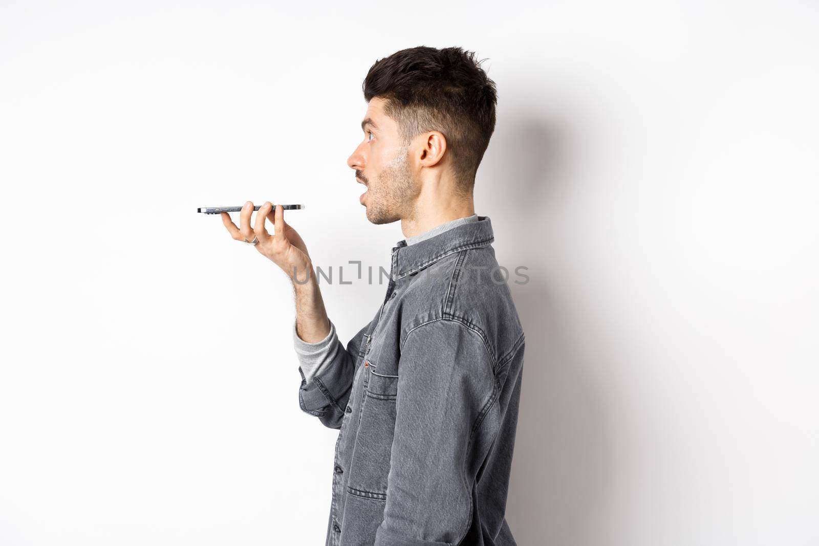 Profile of young man speaking at smartphone, translate with voice-translator app, talking into mobile phone speakerphone, standing on white background.