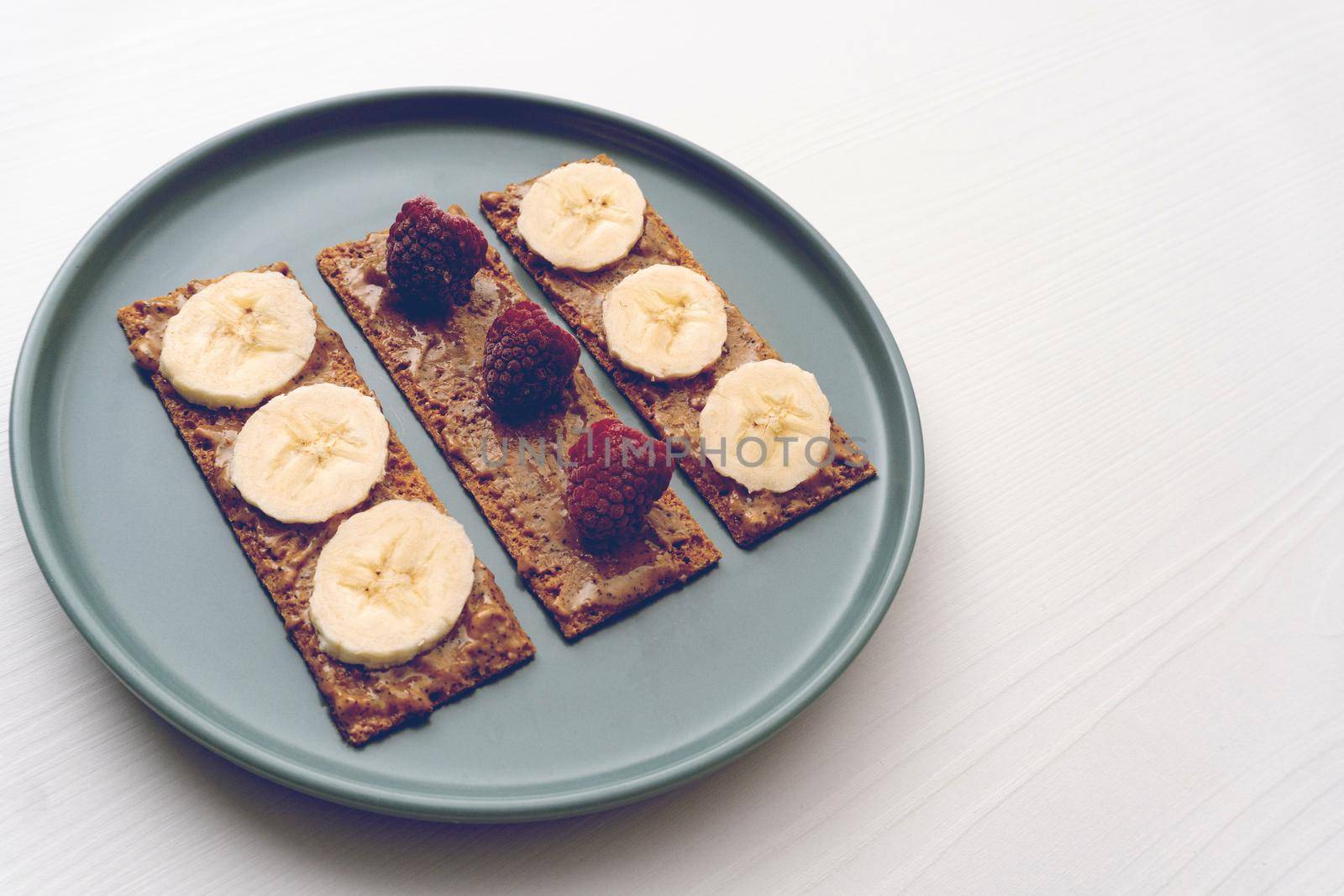 Peanut butter, raspberry and banana on bread slices on round plane on white table, toast for morning breakfast or snack for brunch, minimal concept, healthy eating, toned