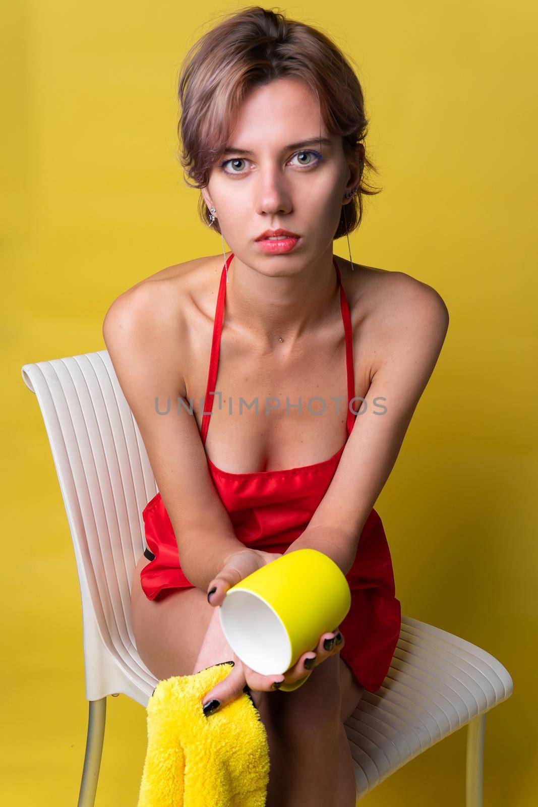 Girl cleans up with a rag and a cup on a yellow background in a red apron housekeeping washing, yellow microfiber house . maid iew portrait protective rubber housewife working holding