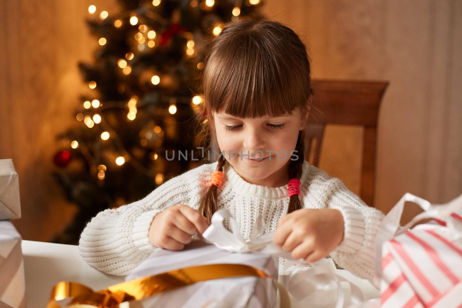 Indoor shot of cute charming little girl wearing white sweater packing presents for her parents and friends, smiling happily while sitting at table in living room with decorated fir tree on background