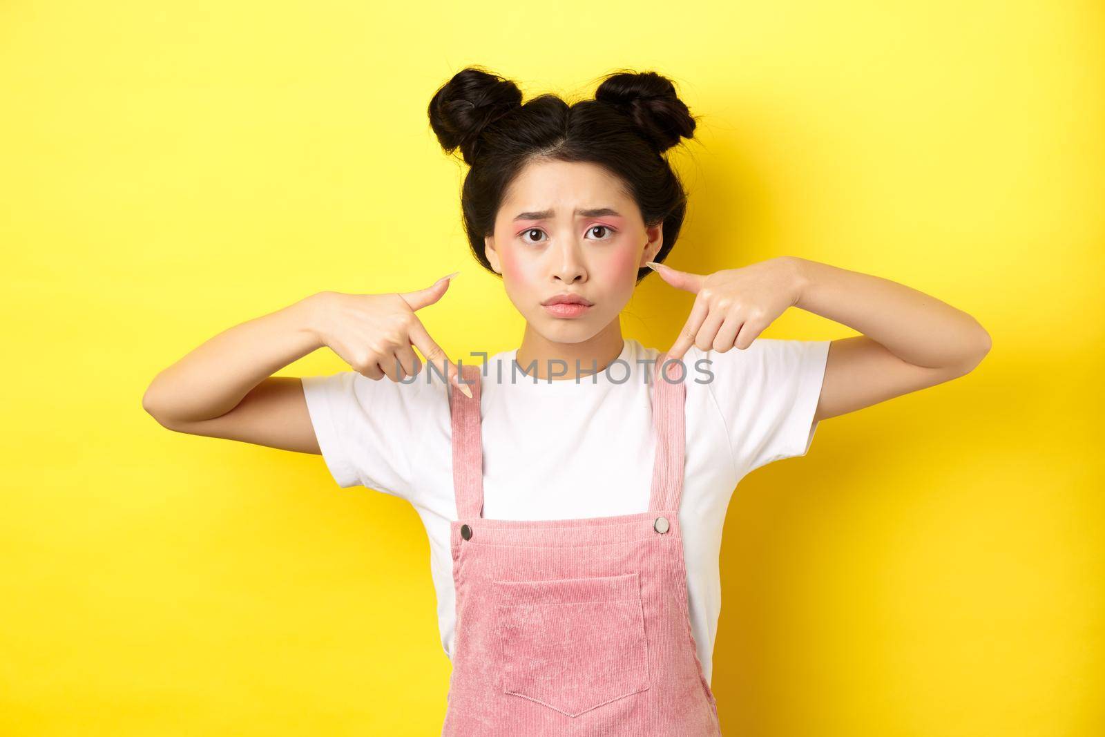 Worried and confused asian girl with glamour makeup, pointing fingers down and look sad, standing in summer clothes against pink background.