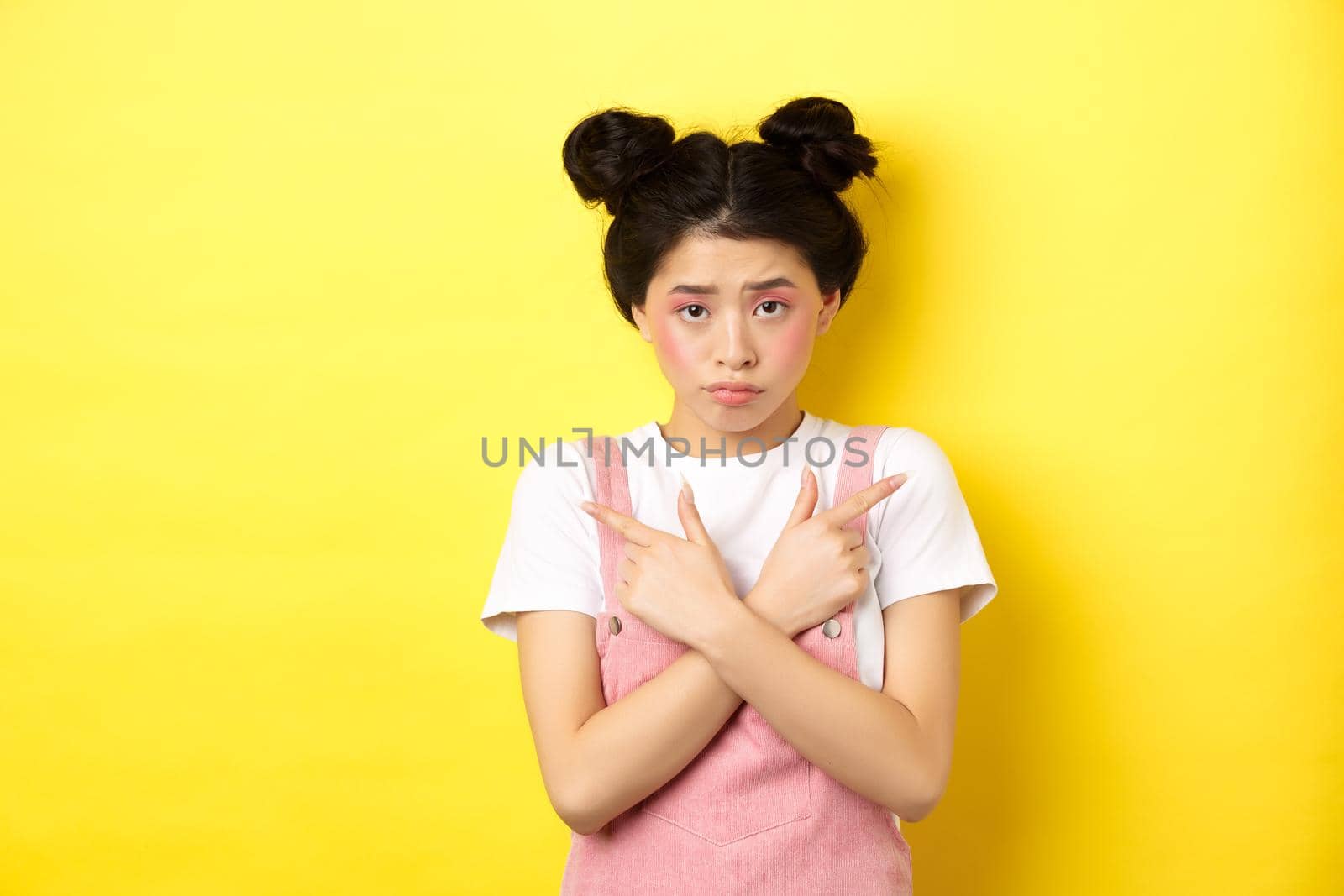 Indecisive timid asian girl pointing sideways, showing left and right ways and sulking upset, standing sad on yellow background by Benzoix