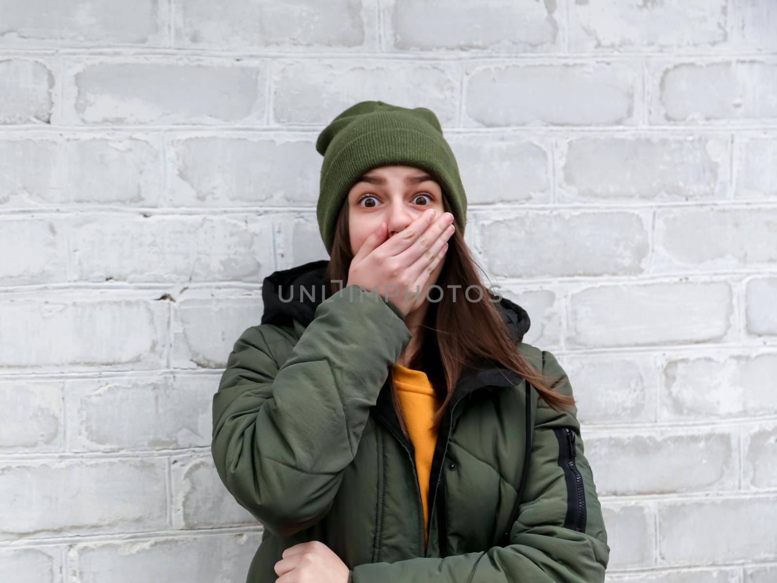 Portrait of a beautiful shocked girl who closes her mouth with her hand in a yellow sweater and khaki hat that stands near a white brick wall. The concept of emotions. by lunarts
