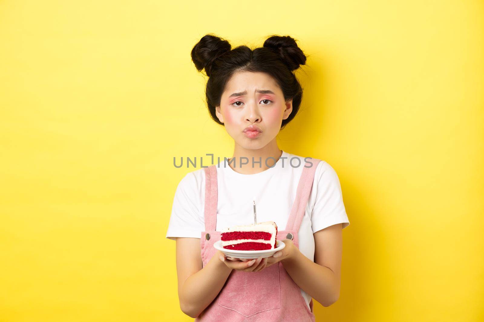 Sad and lonely birthday girl frowning upset, holding birthday cake with candle, making wish, standing on yellow background.