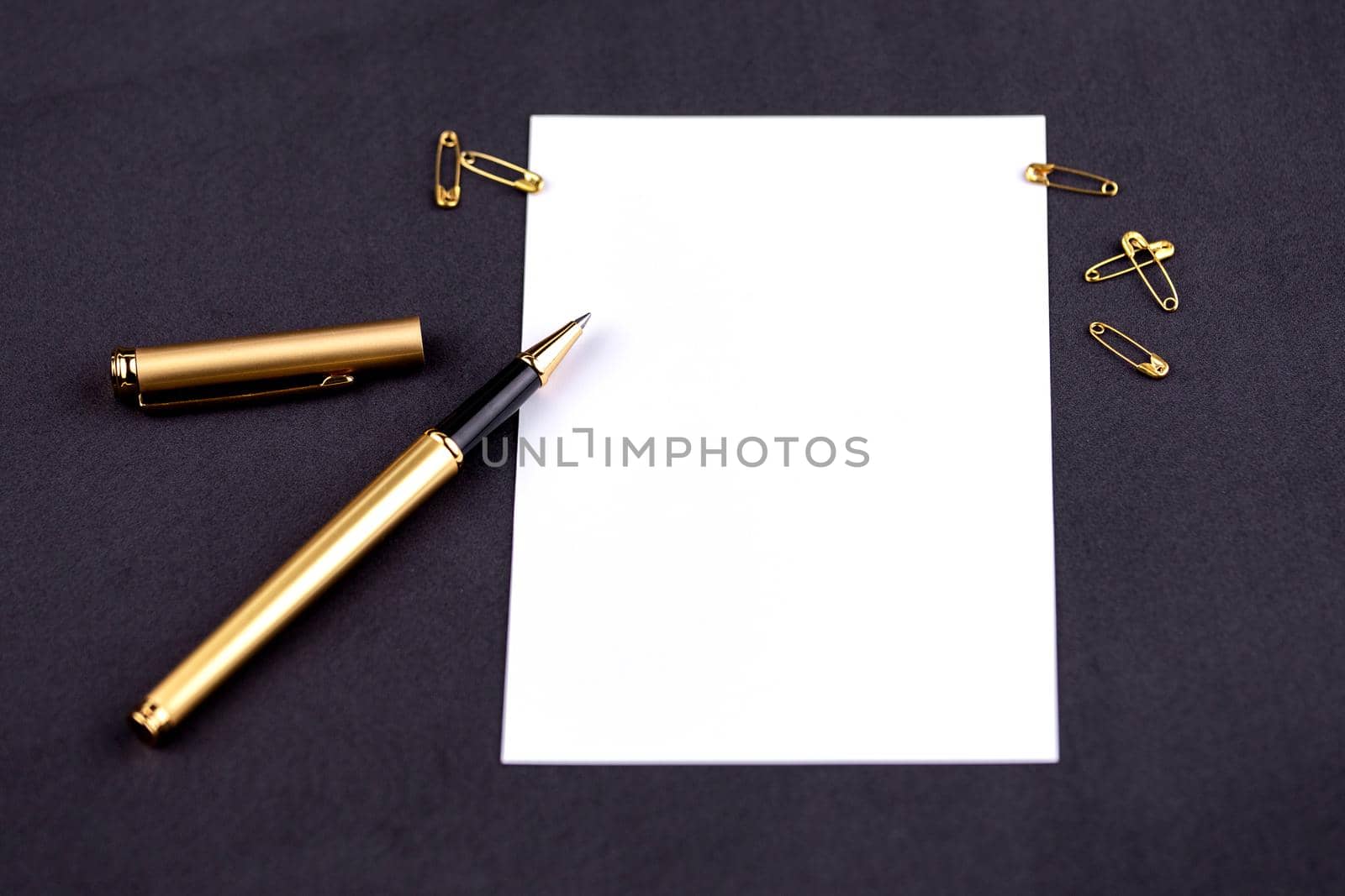 Gold pen, ribbon, paper clips and stationery on a black background with a white sheet of paper.