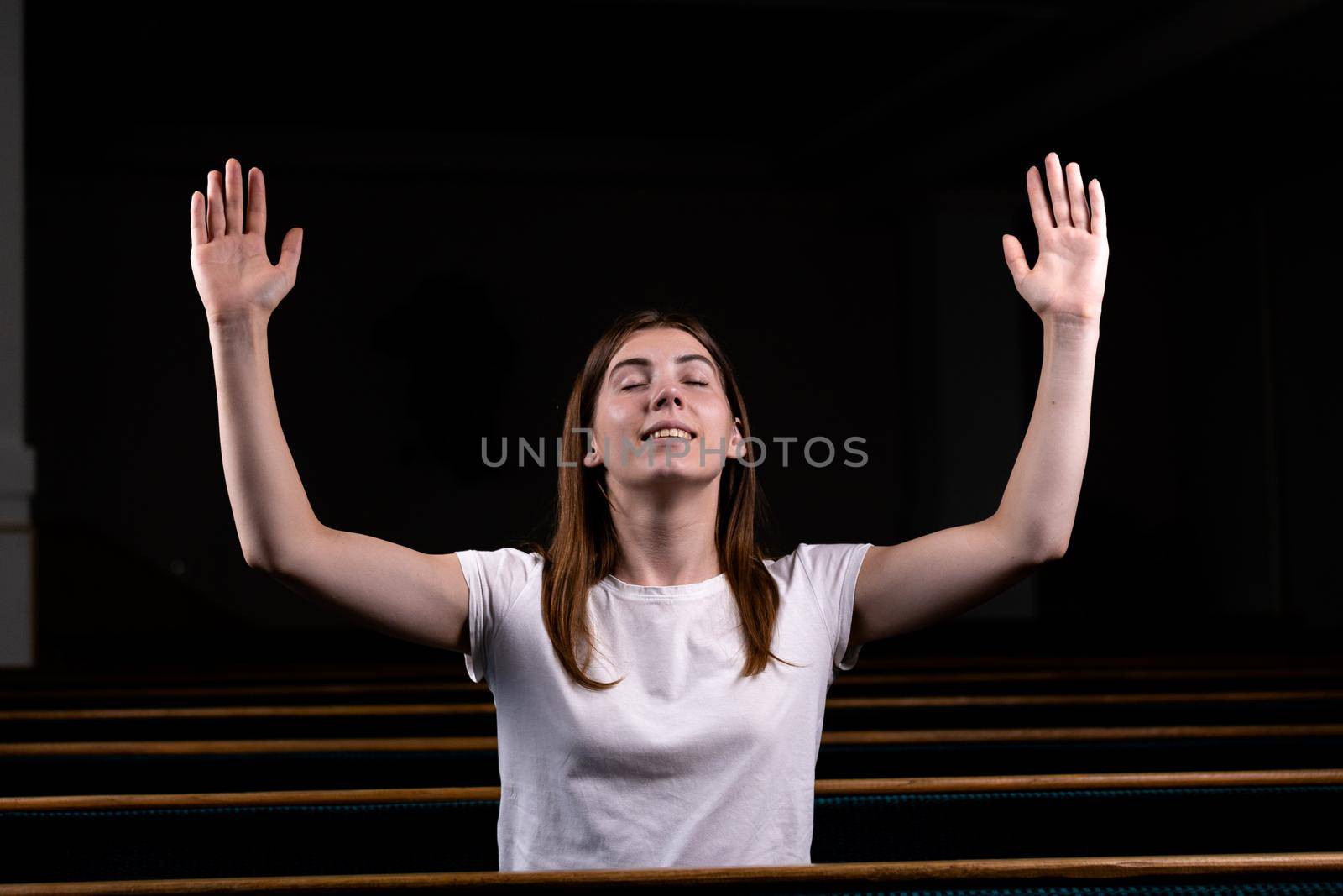 A Christian girl in white shirt is sits with his hands up and face and praying with humble heart in the church.