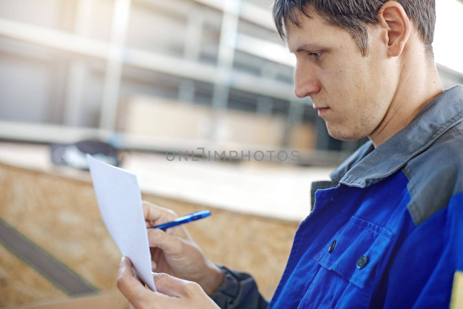 A warehouse worker in blue overalls accepts and describes the goods in the room. A young man of Caucasian appearance takes the goods in the warehouse and makes entries in the magazine. Attentive and focused employee.