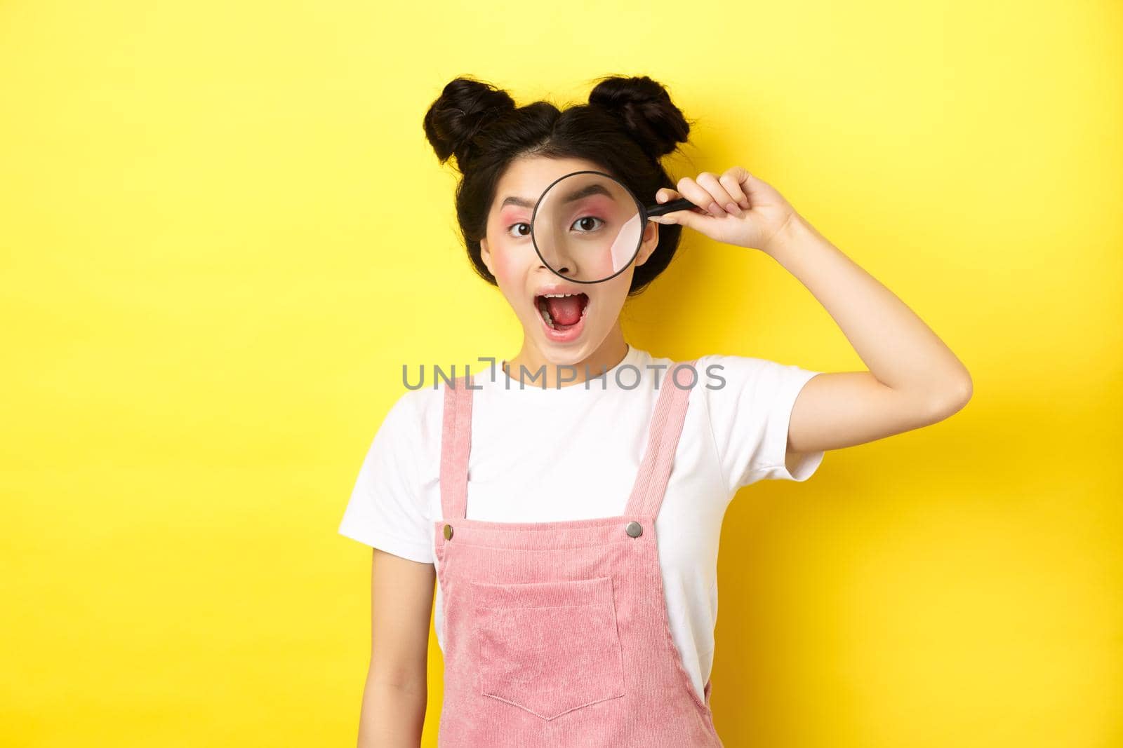 Young asian girl with bright makeup look through magnifying glass, smiling excited, seeing interesting promo, yellow background.