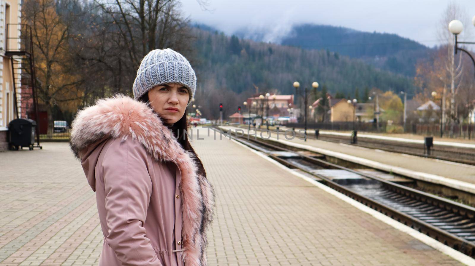 A woman in a gray knitted hat and a pink jacket with fur is waiting for the train at the station looking at the arriving train. Rail transportation. Autumn and winter travel concept