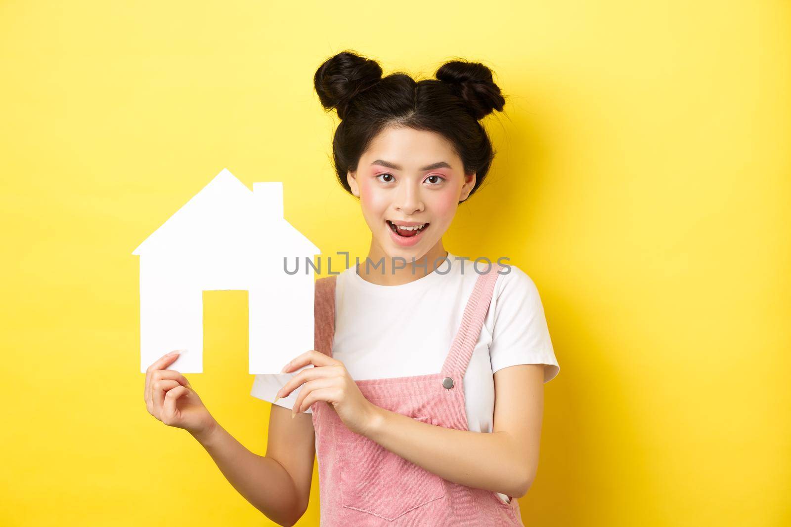 Real estate and family concept. Cute asian woman with bright makeup and stylish hairbuns, showing paper house cutout, smiling determined, yellow background by Benzoix