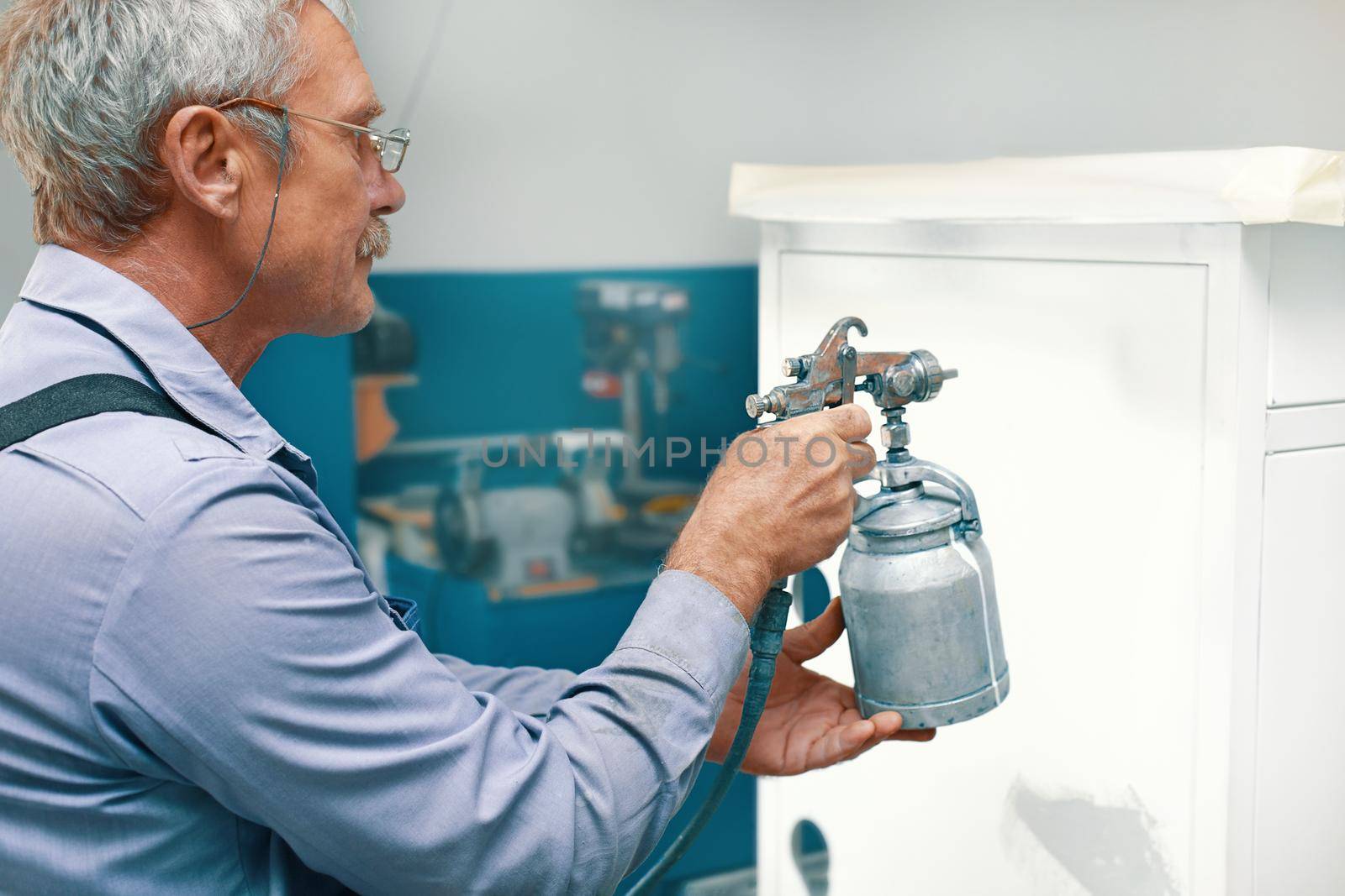 An employee paints metal products. An elderly man holds a compressor spray gun in his hand and paints the Cabinet white