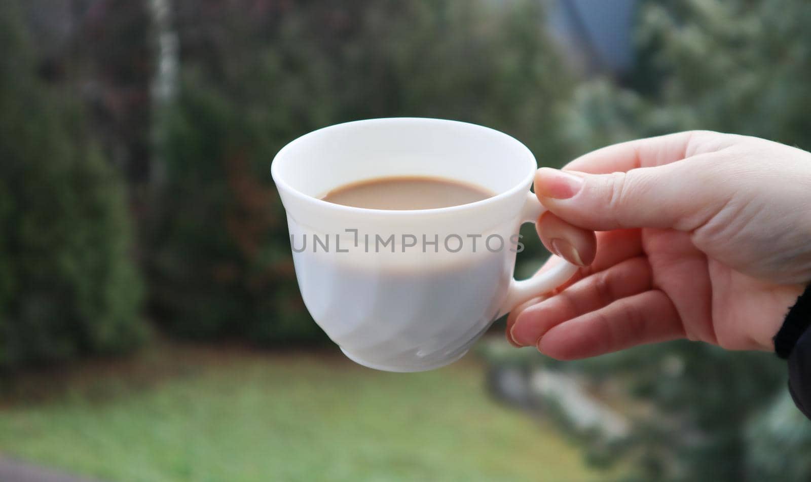 Girl hand holds a cup of white coffee and has a blurred green nature background. Mug in a female hand on a background of mountains