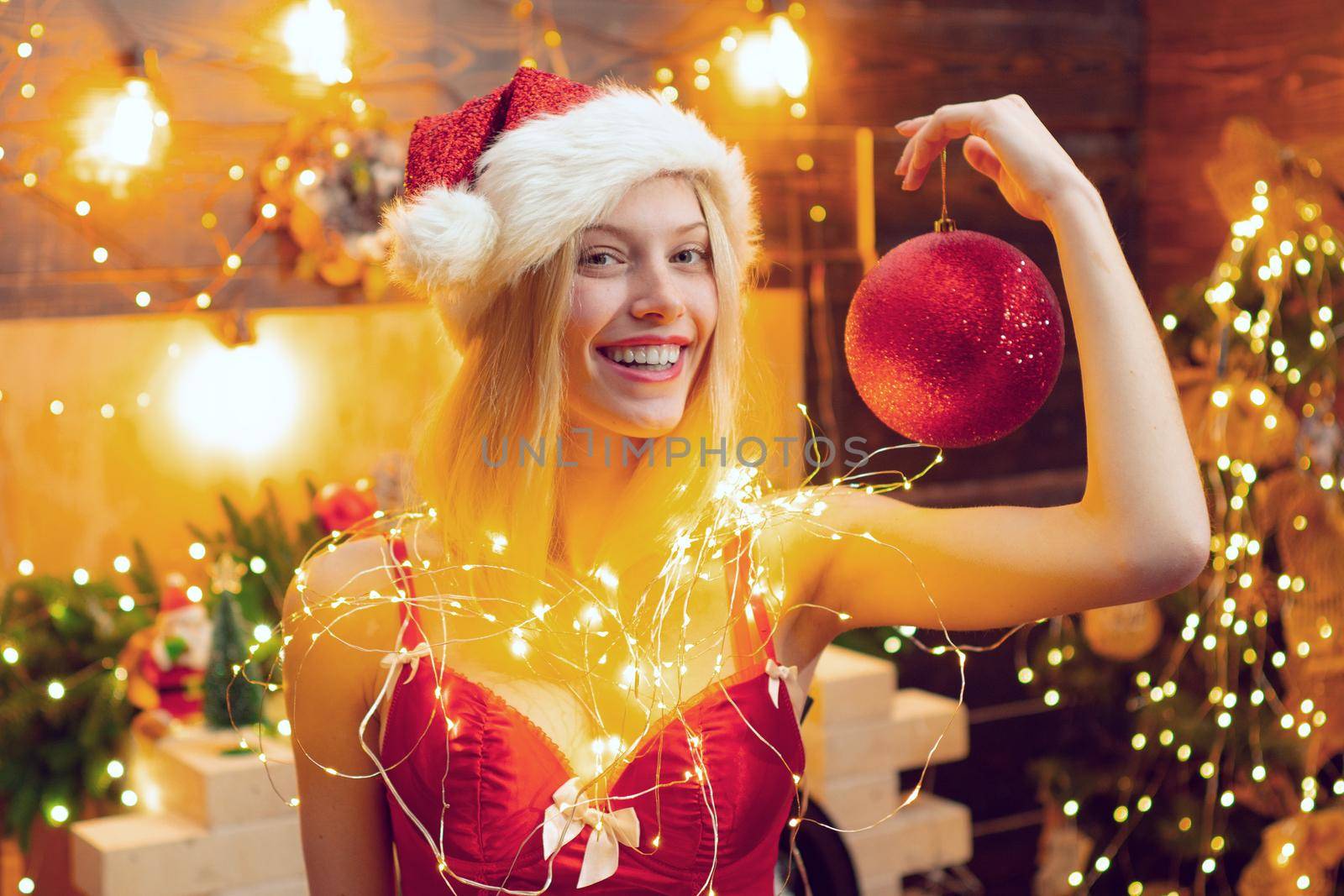 Smiling woman with red lips in christmas light is looking at camera. Girl is wearing Santa hat. Concept of holidays. by Tverdokhlib