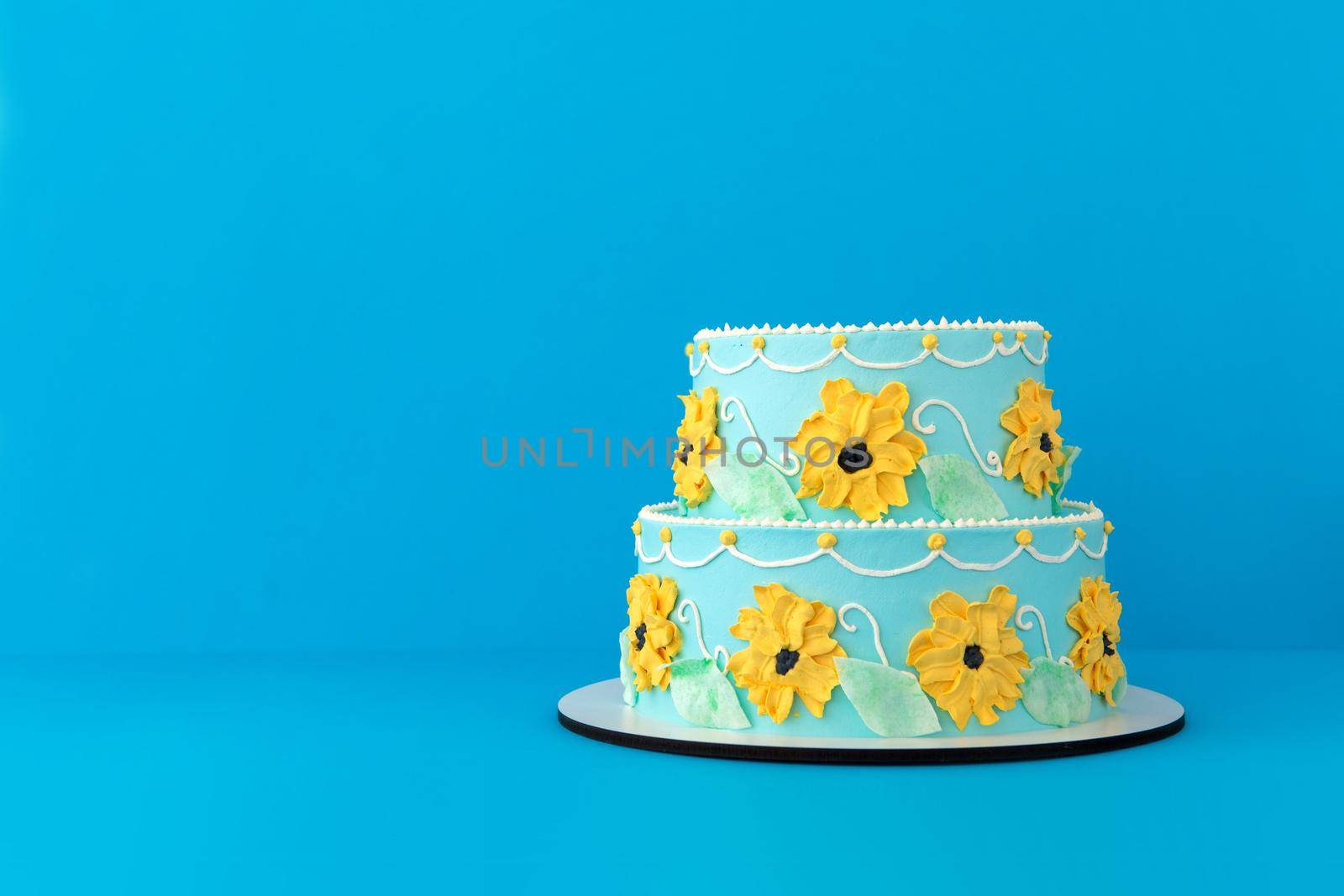 Colorful tiered festive birthday cake decorated with yellow flowers and green leaves made of cream on bright blue background