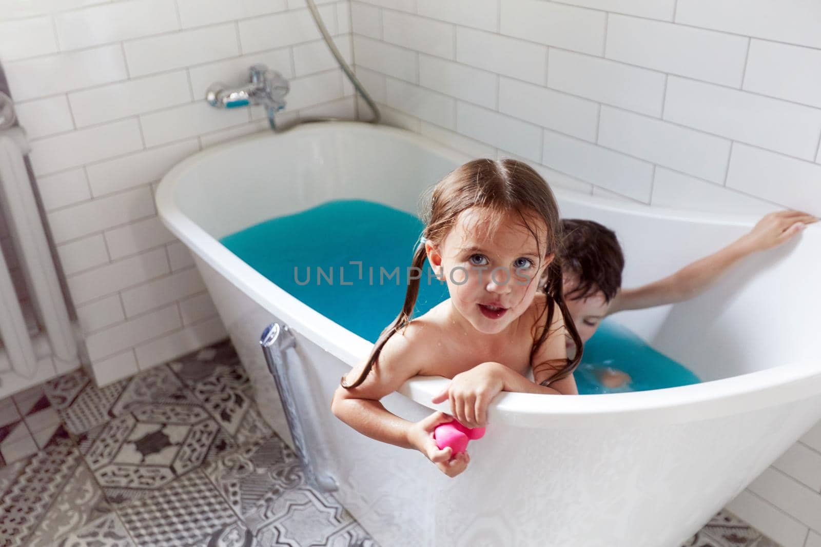From above of adorable little brother and sister bathing in bathtub with blue water and having fun together