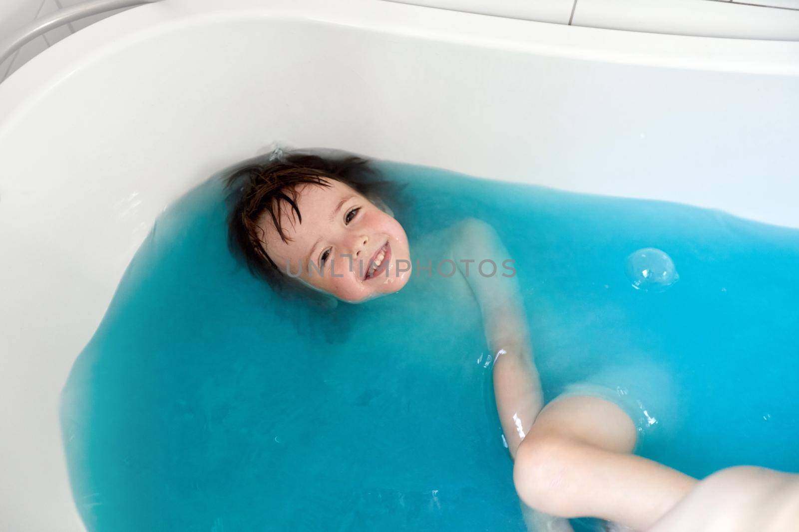 Baby boy swims on his back in blue water in bathtub with tooth smile. Top view of adorable little kid laughs relaxing during hygiene routine