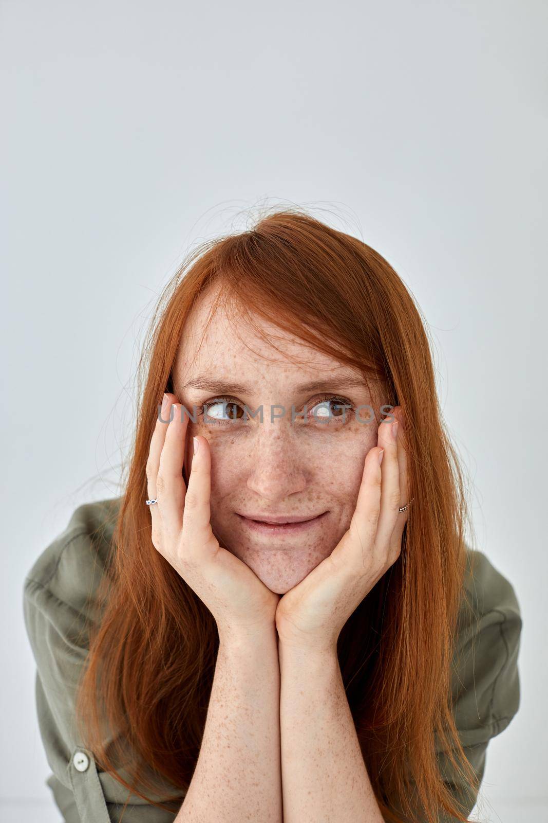 Positive female with ginger hair keeping hands on cheeks and looking away against white background