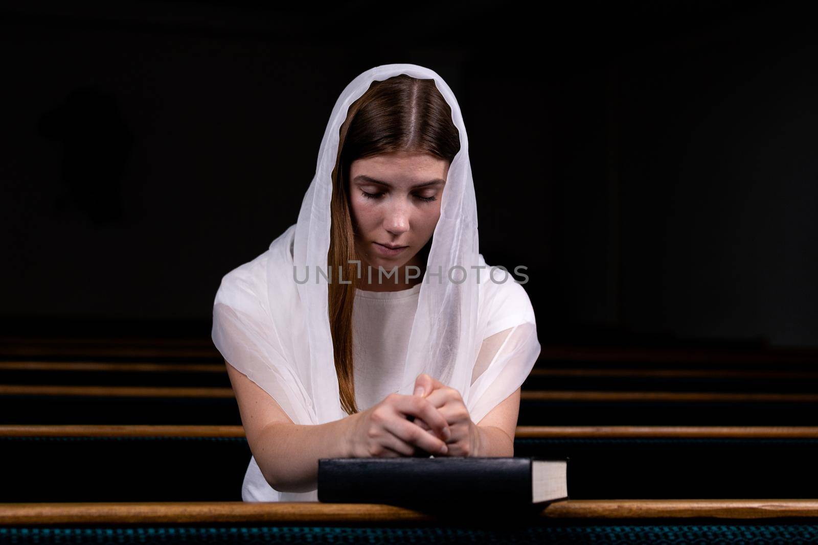 A young modest girl with a handkerchief on her head and a bible in her hands is sitting in church and praying. The concept of religion, prayer, worship.