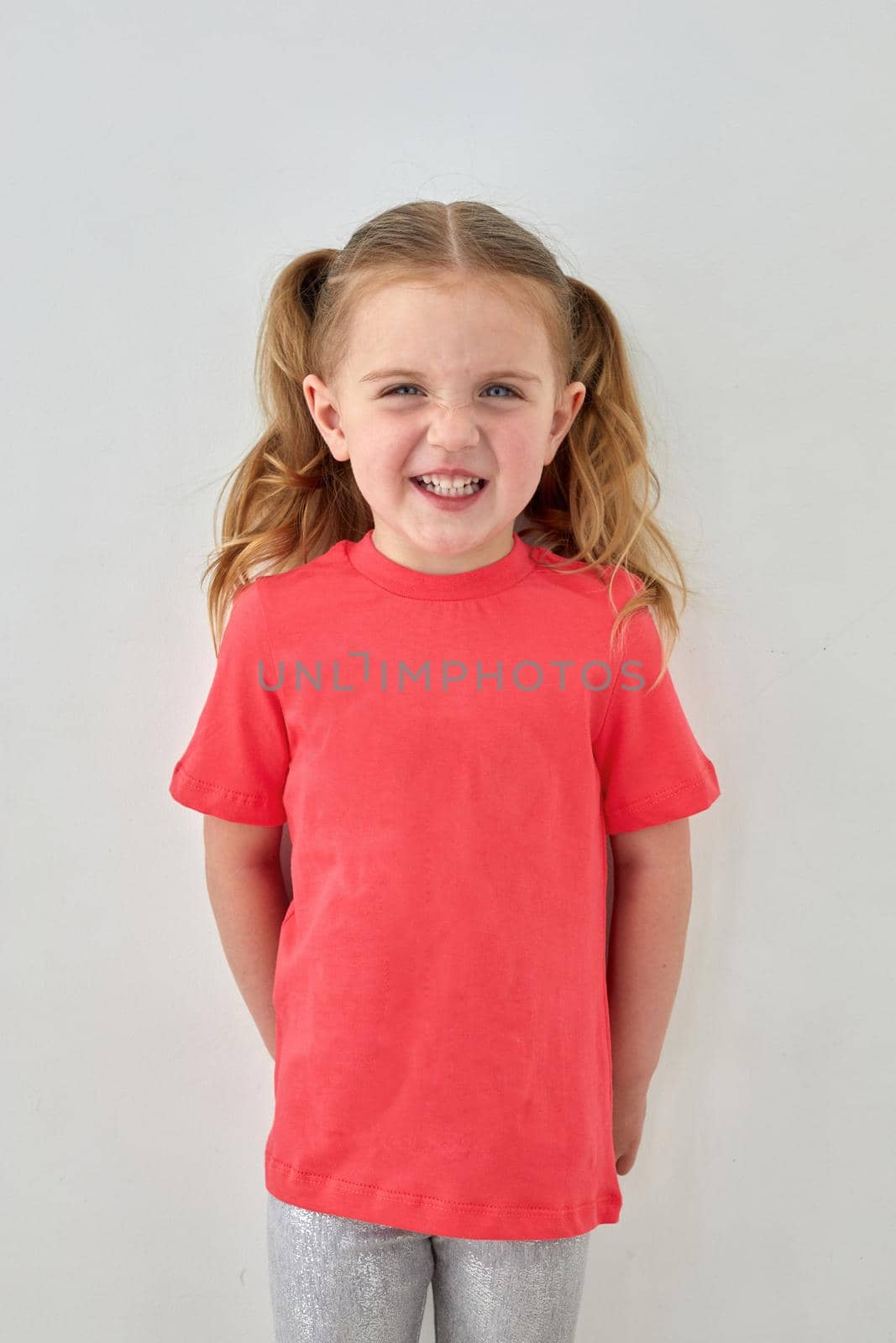 Adorable cheerful little girl with ponytails wearing pink t shirt standing on white background and looking at camera with smile