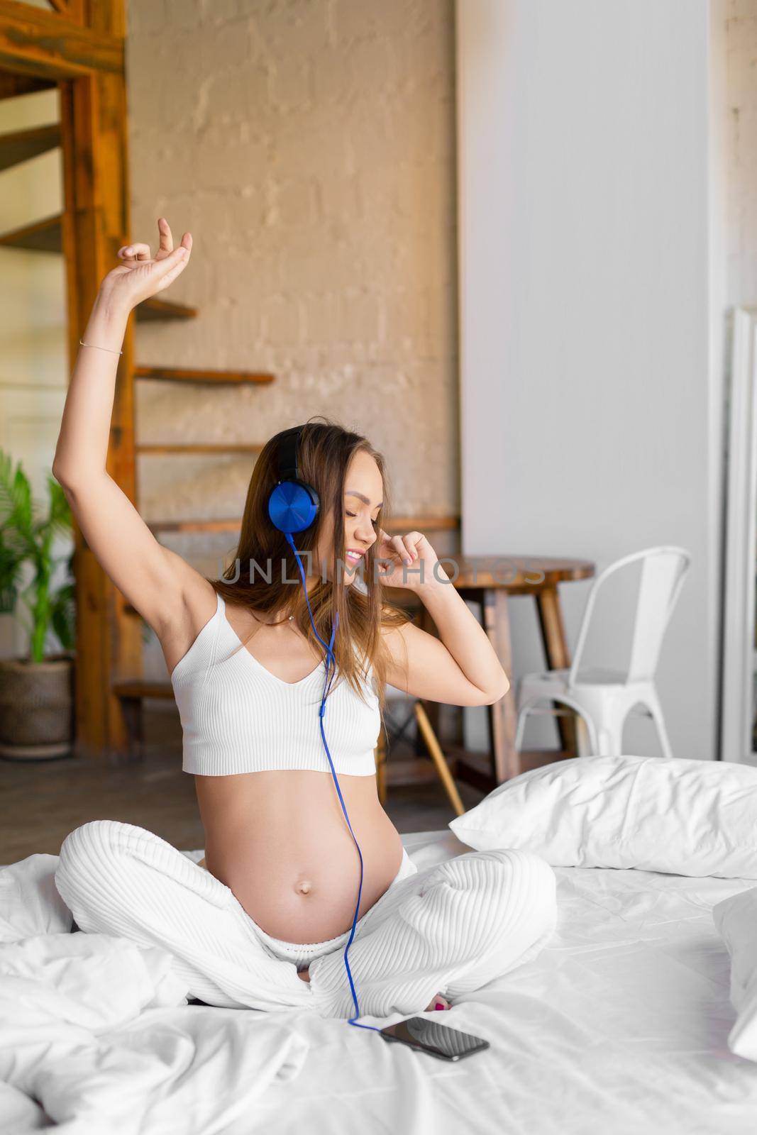 Pregnant woman sits on a bed in home interior and enjoys soothing music. She listening to the music via online smartphone application.