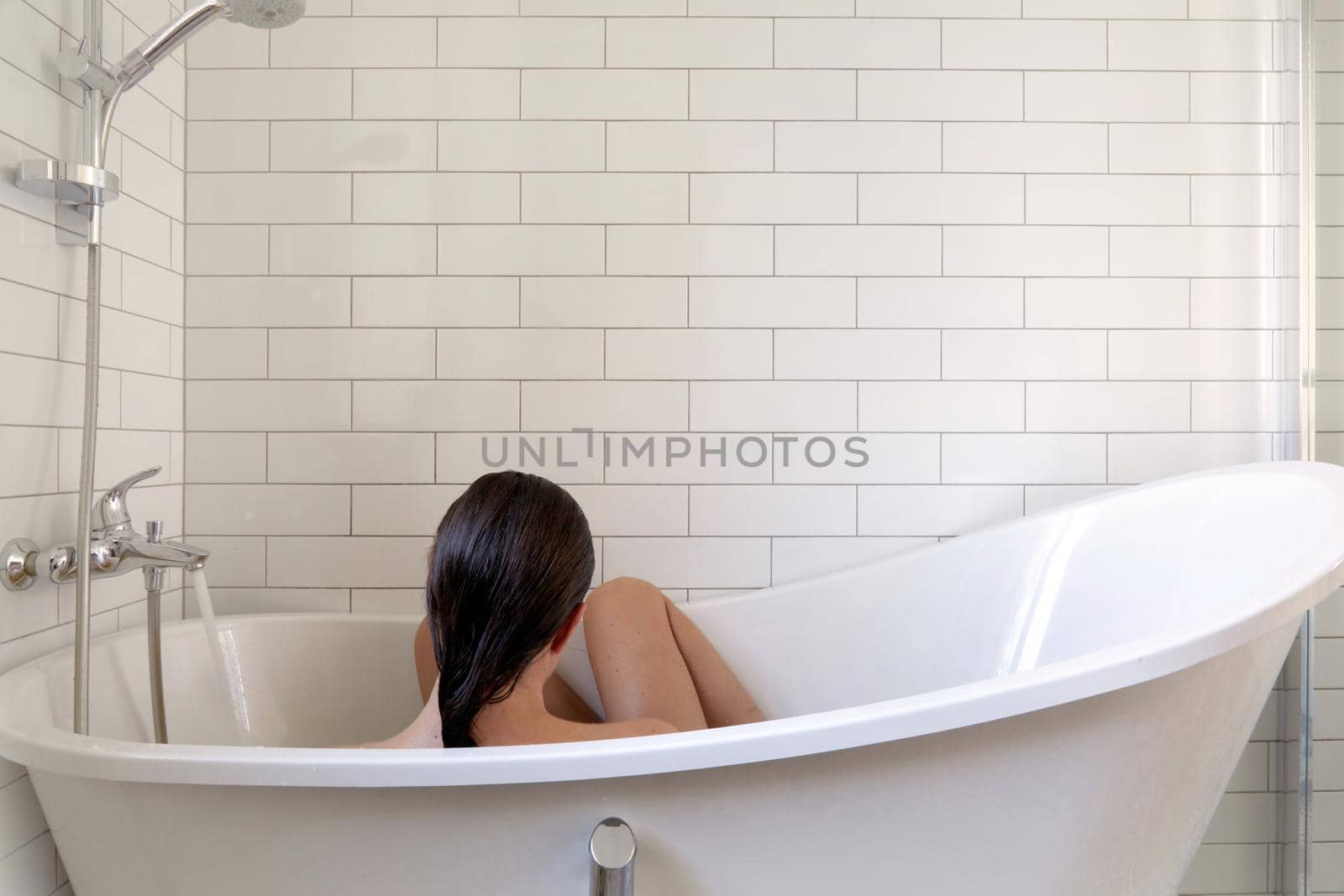 Back view of unrecognizable female with wet hair and skin sitting in bathtub during daily hygienic and skincare procedure