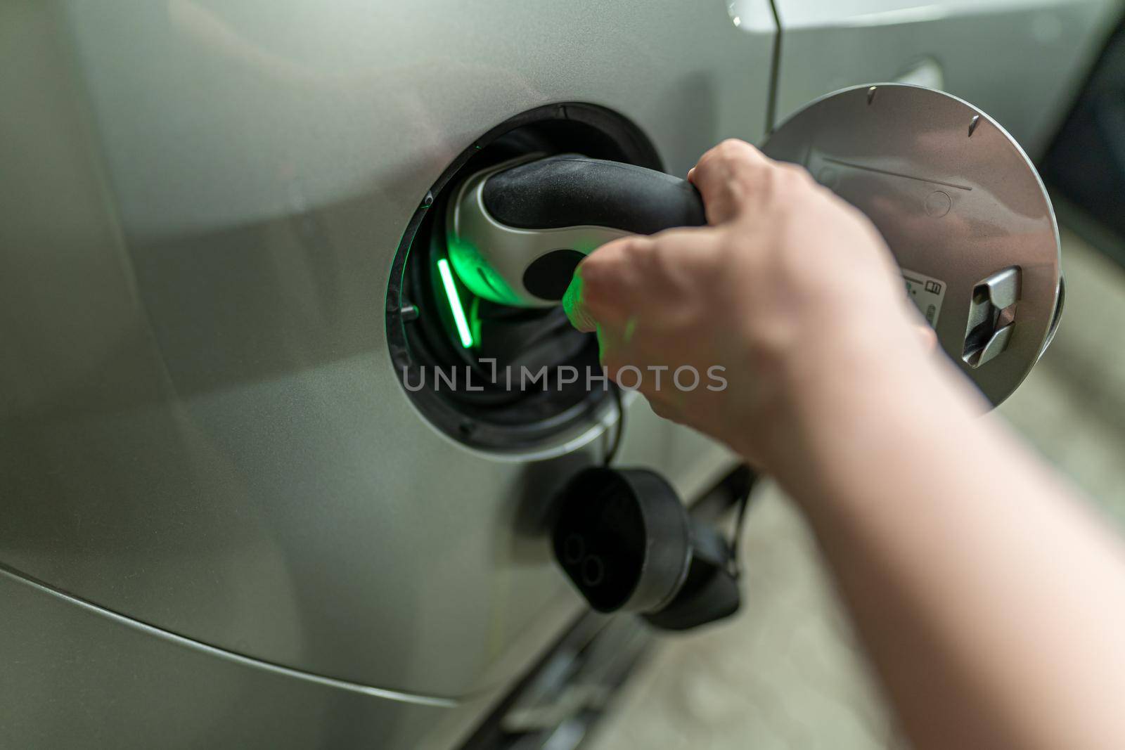 Detail view at a mans hand with is plugin the electric charging wire and plug into his e-automobiles fuel cap. Green light for the active charhing process