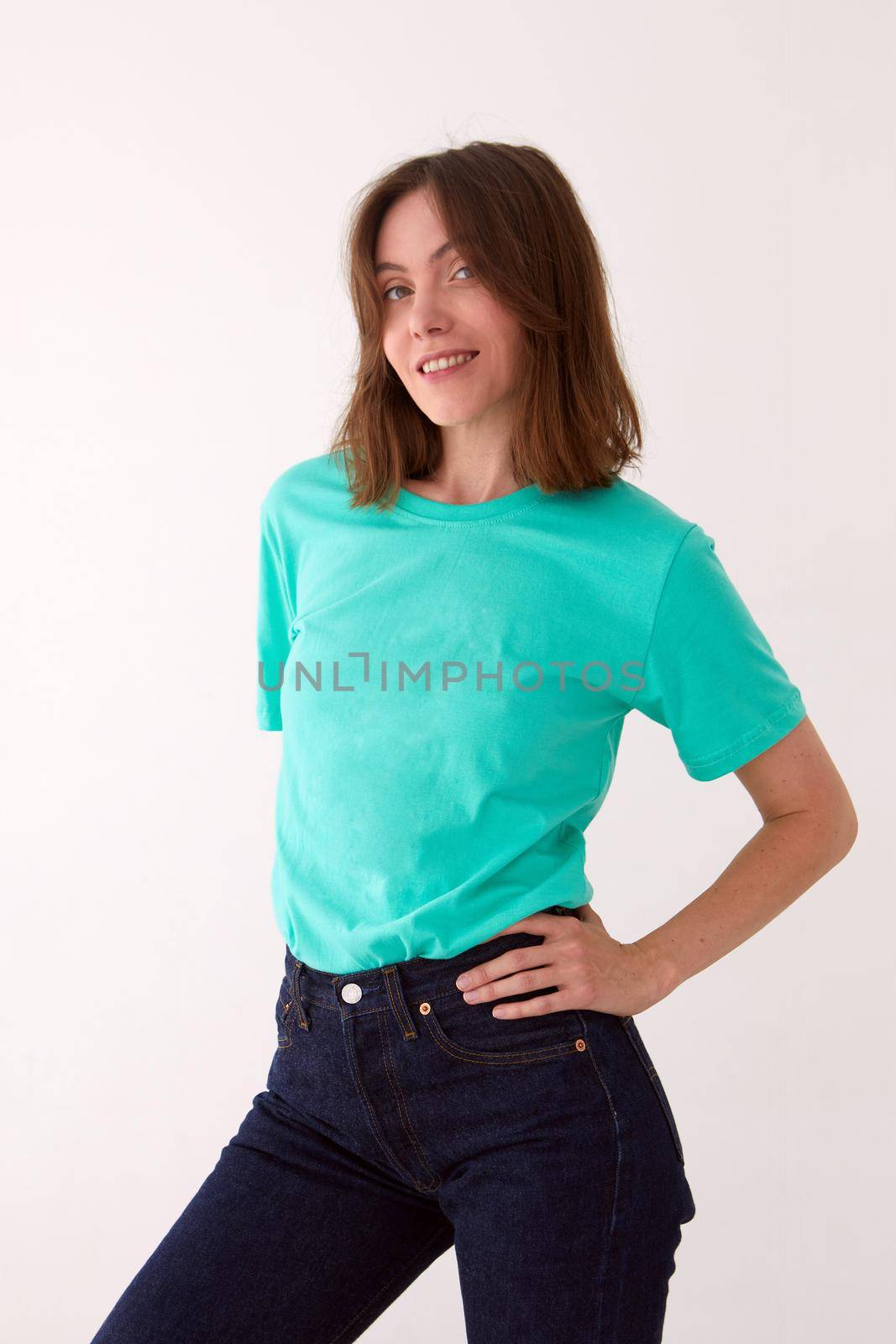 Positive female model wearing t shirt and jeans standing with hands on waist against white background and looking at camera