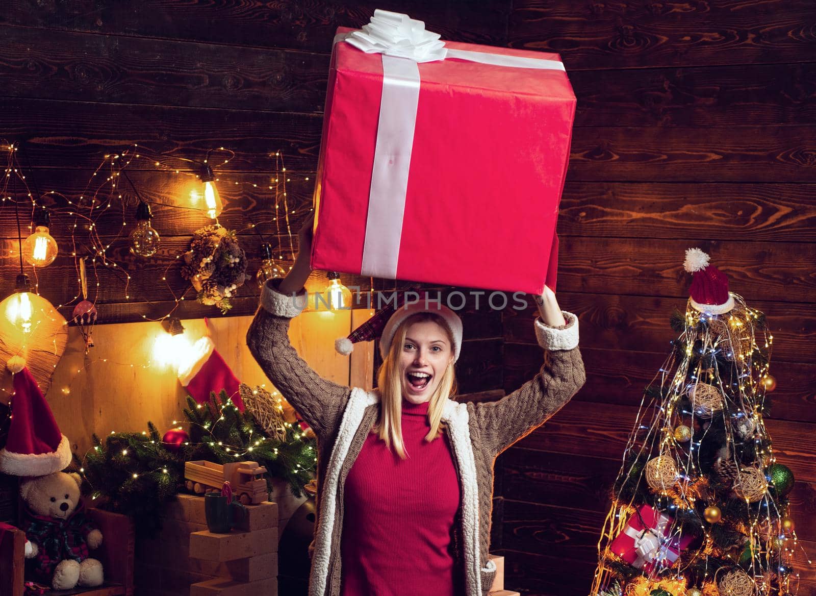 Christmas woman with gift in her hands under her head. Happy young lady by the fireplace near the Christmas tree. Excited blonde girl with big gift box