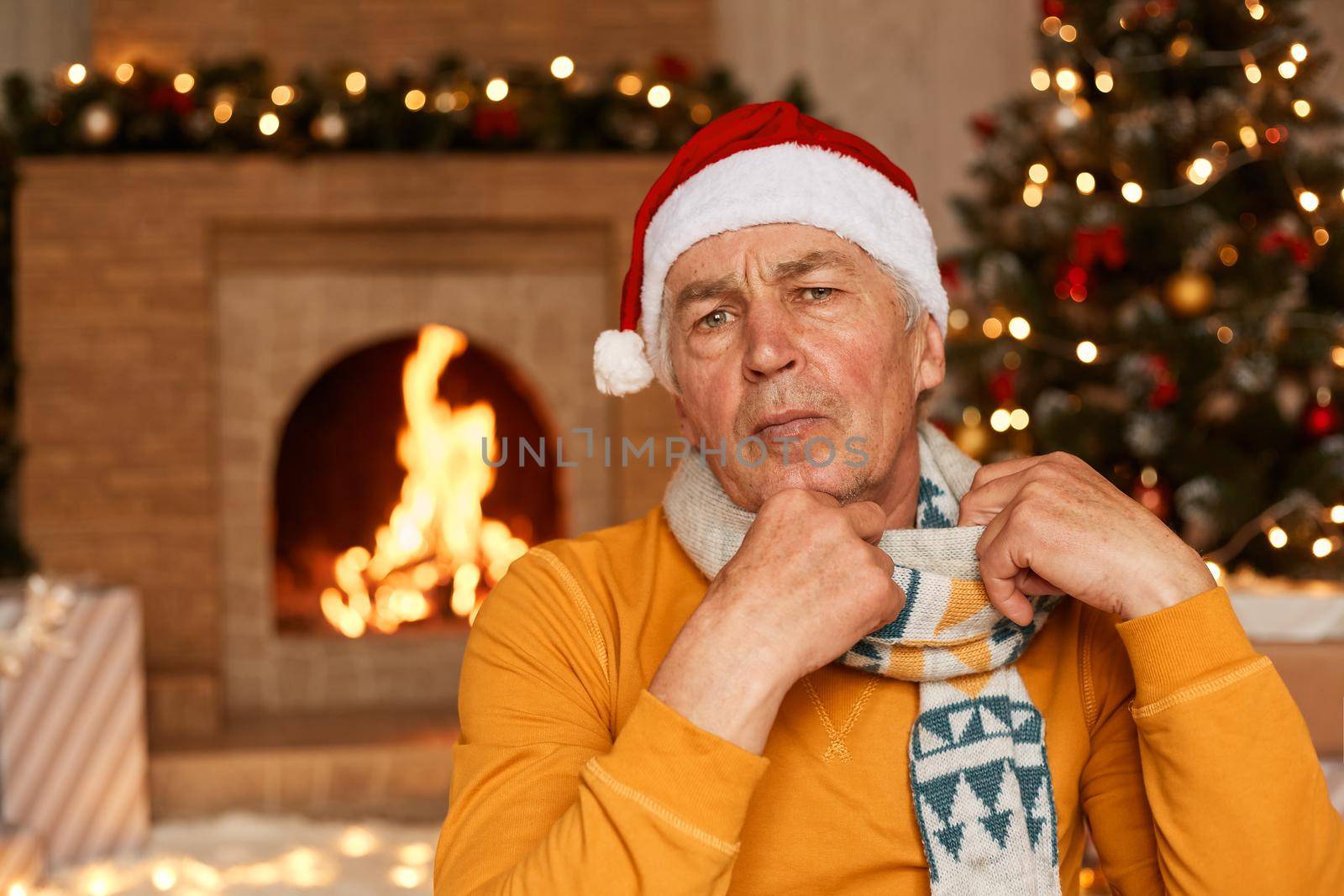 Portrait of mature man wearing orange sweater, scarf and santa claus hat posing in living room at home near christmas tree and fireplace, looking at camera, celebrating new year alone. by sementsovalesia