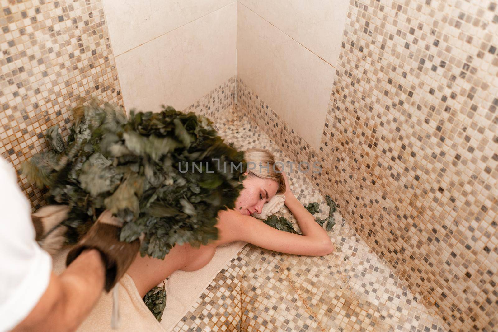 Slim woman in bath is getting steaming massage with hot oak leaves brooms, in steam-room.