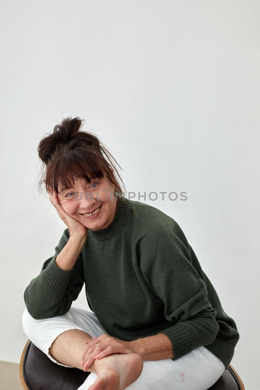 62 years old woman props her head with her hand and looks at the camera with a grin against a white wall background. Woman keep her head on hand
