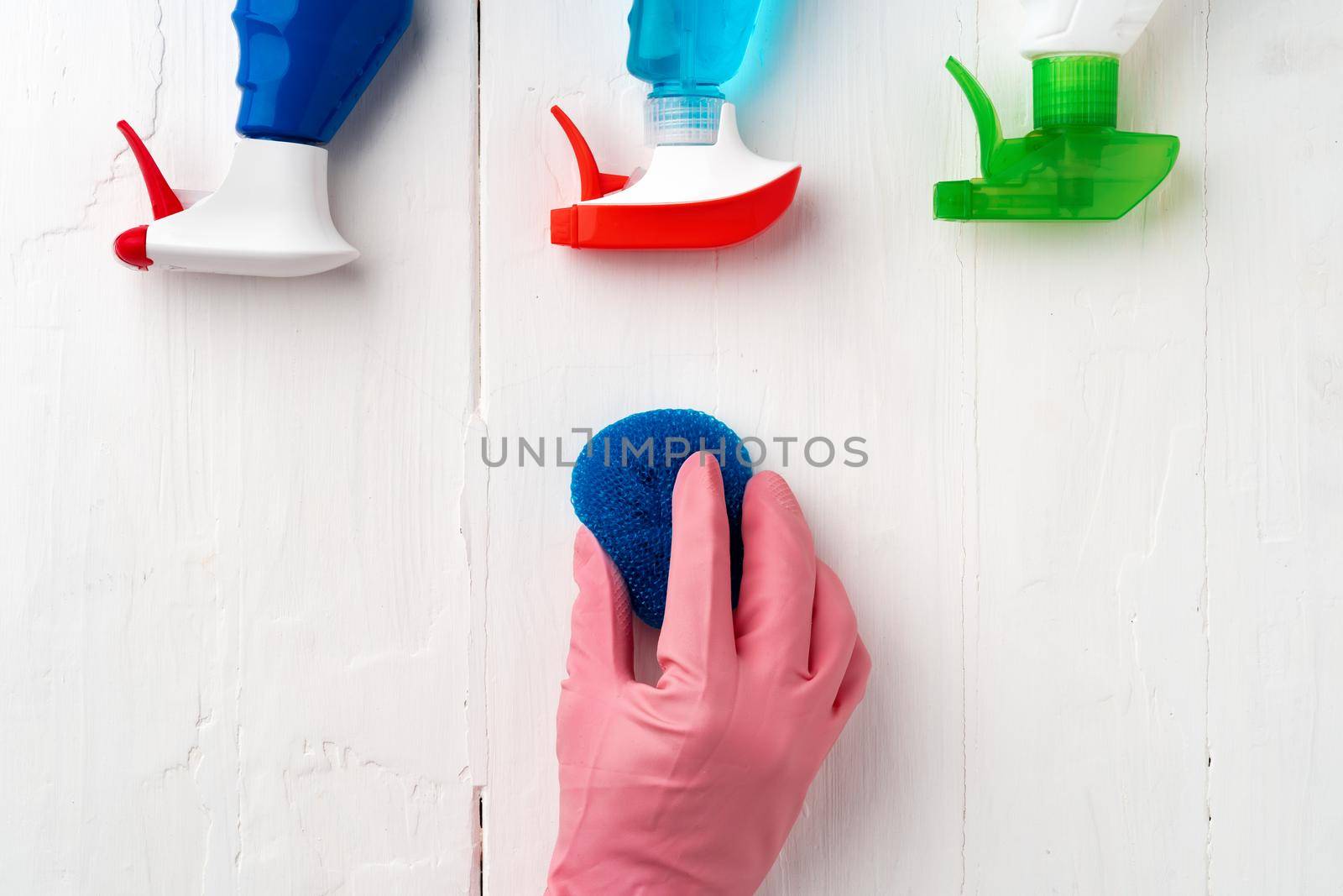 Hand in glove with cleaning supplies above white wooden background, top view