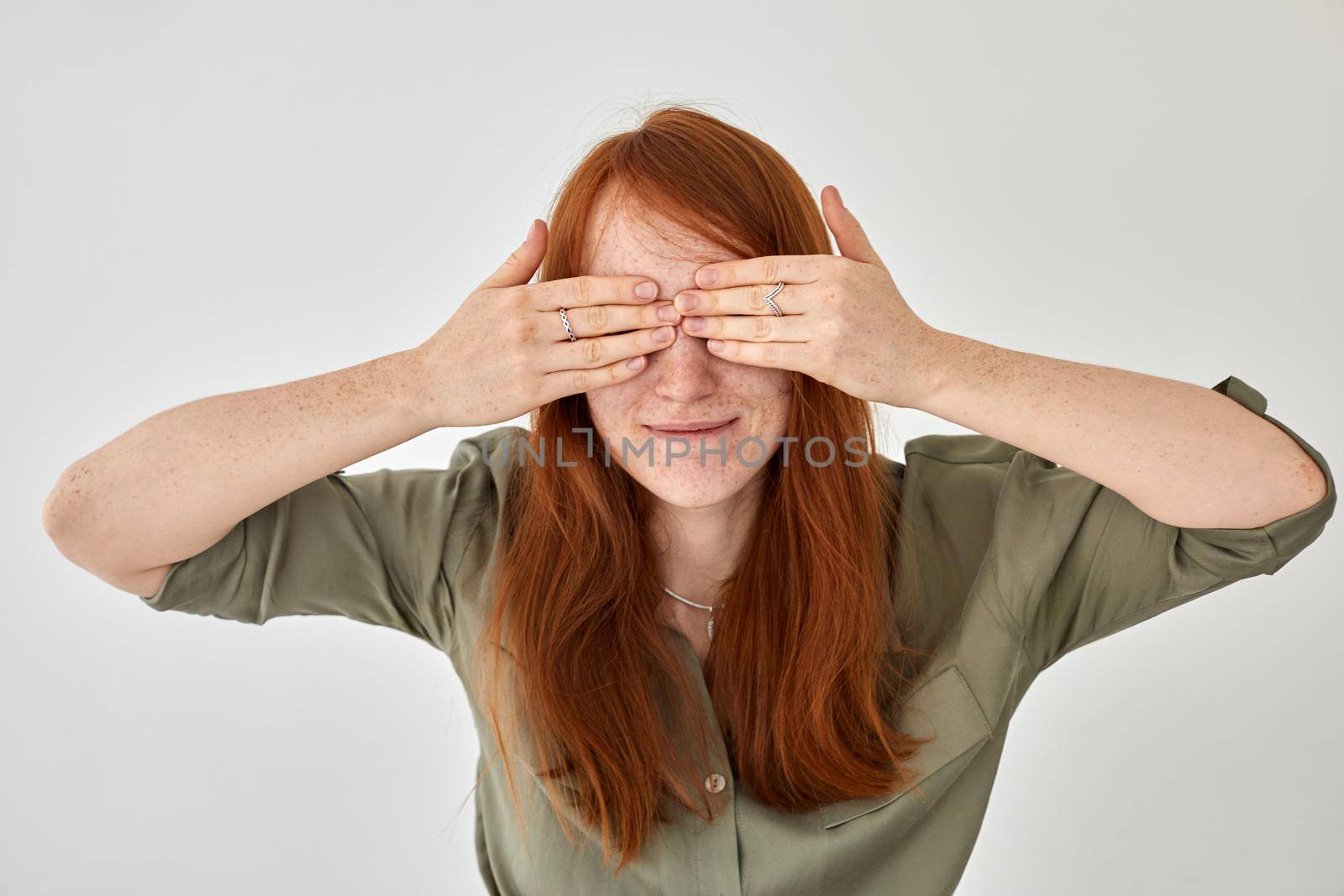 Glad young female with long ginger hair and freckles covering eyes with hands while standing on white background in studio