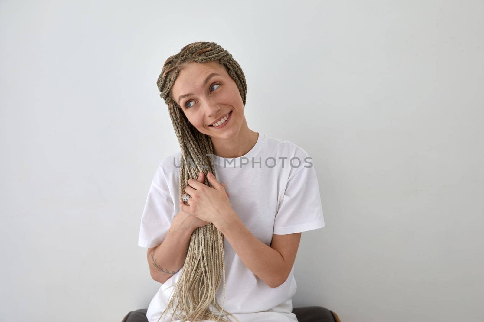 Positive female wearing white t shirt sitting near wall and touching braids while looking away