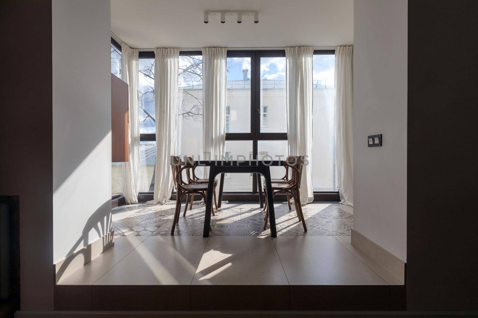 Dining table and chairs placed in front of panoramic window decorated with white curtains in modern light apartment with minimalist interior design