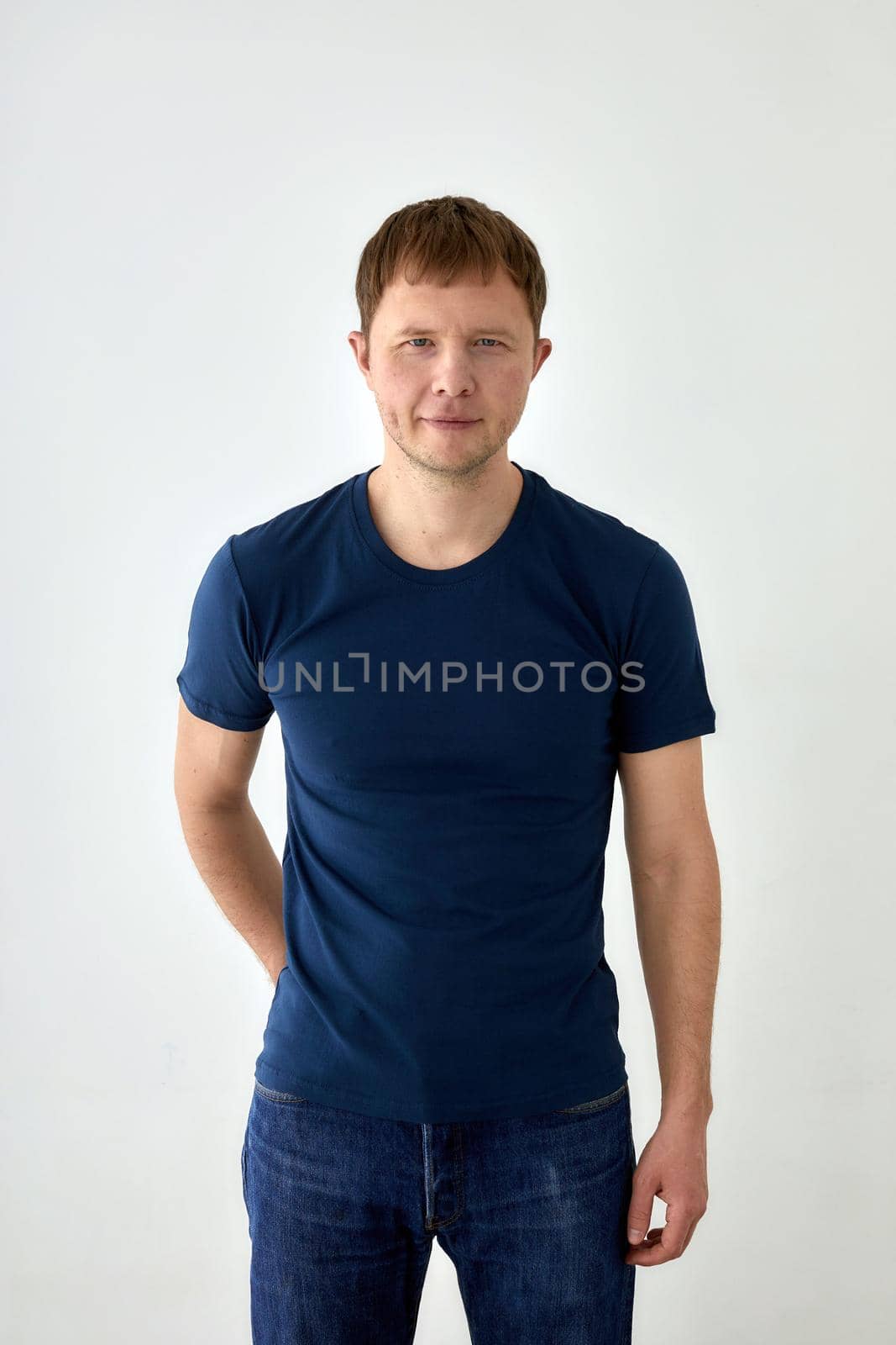 Positive young male in jeans with hand behind and dark blue t shirt standing against white background and looking at camera