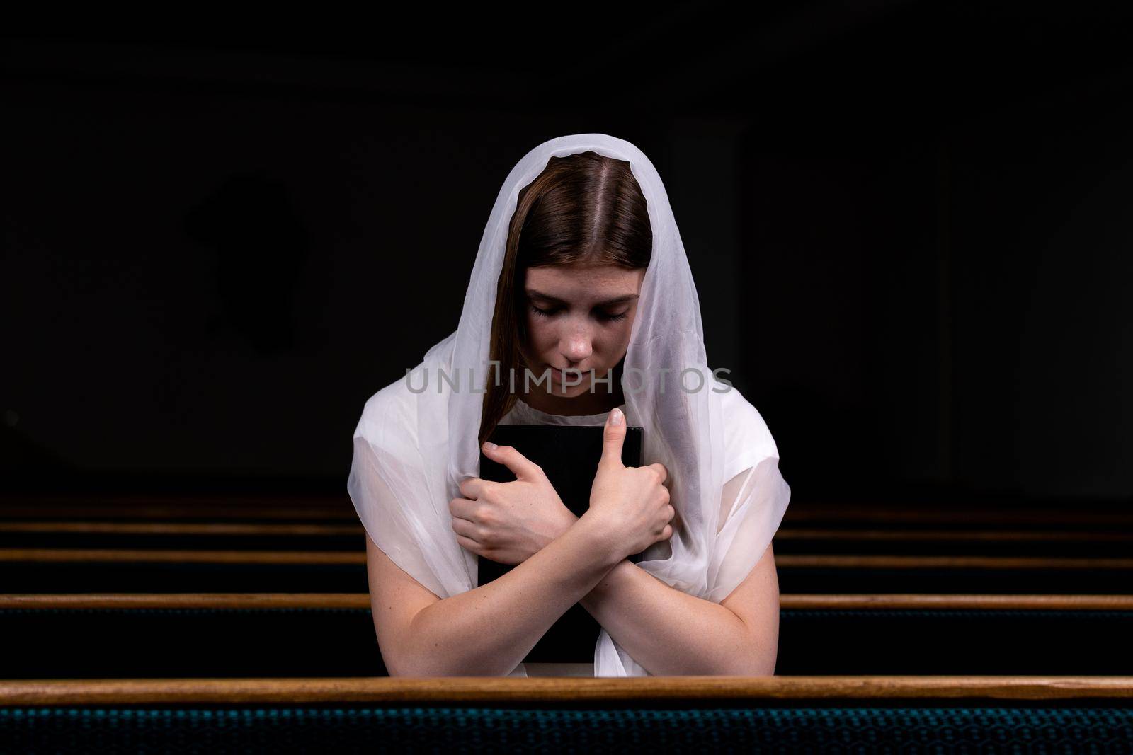 A young modest girl with a handkerchief on her head and a bible in her hands is sitting in church and praying. The concept of religion, prayer, worship.