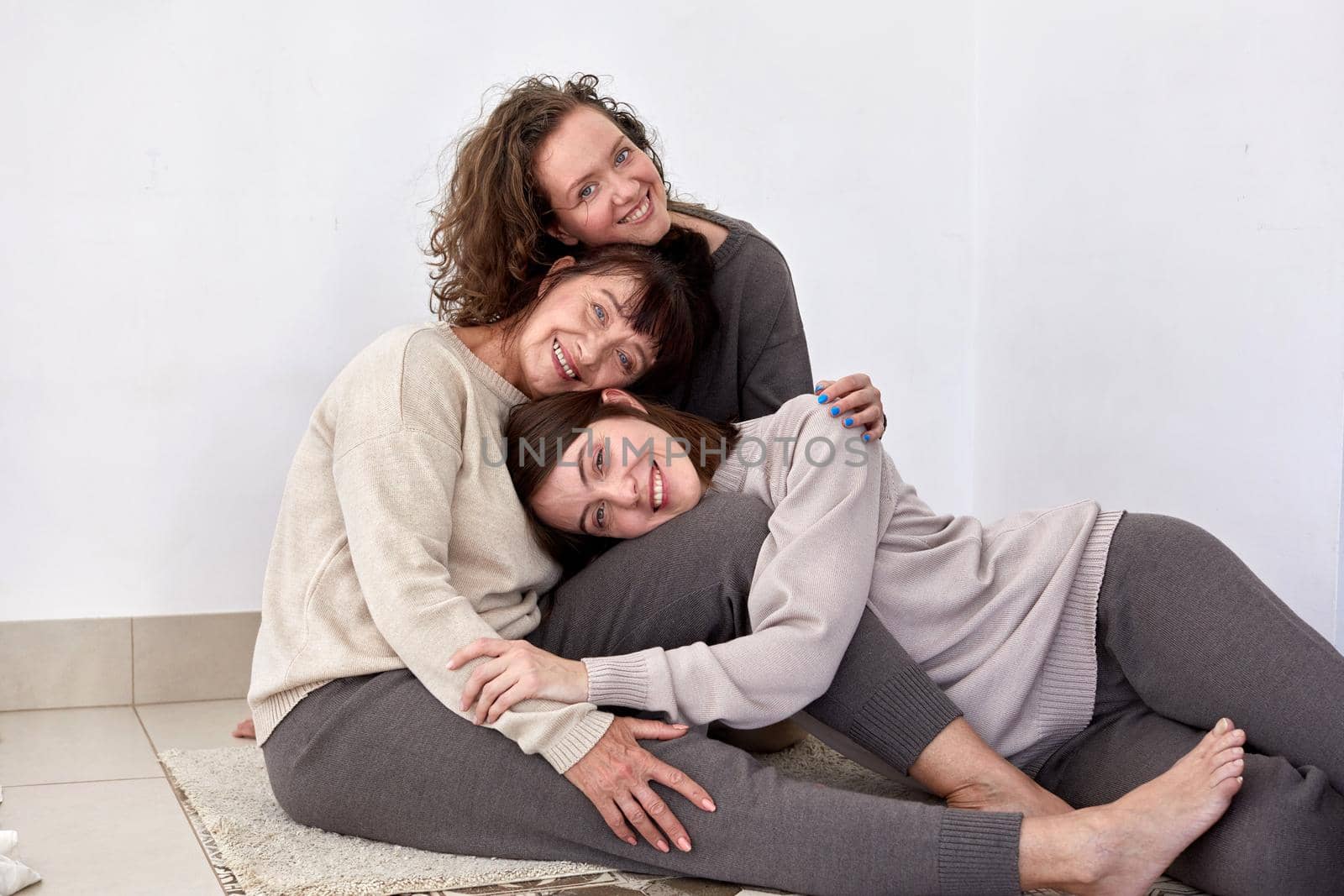 Cheerful adult mother embracing charming young daughters while lie pyramid head on head on white background in studio and looking at camera