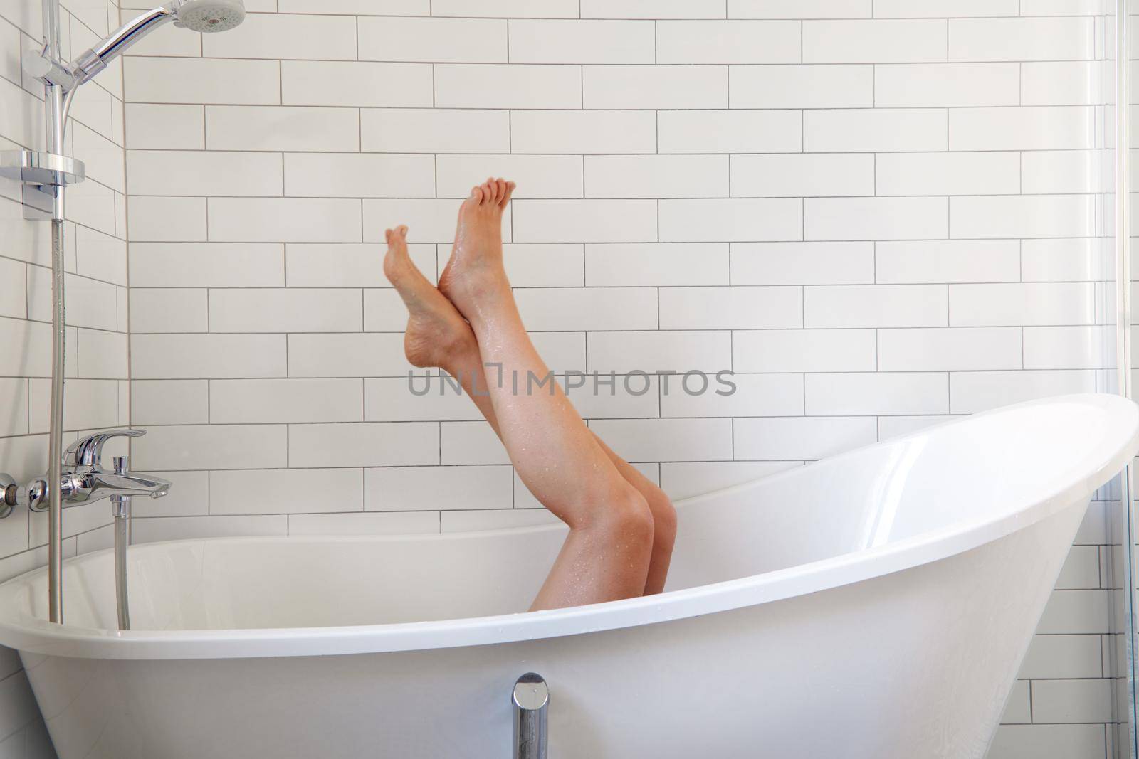 Unrecognizable barefoot female lying in bathtub and raising legs against white tiled wall during skin care routine in bathroom at home