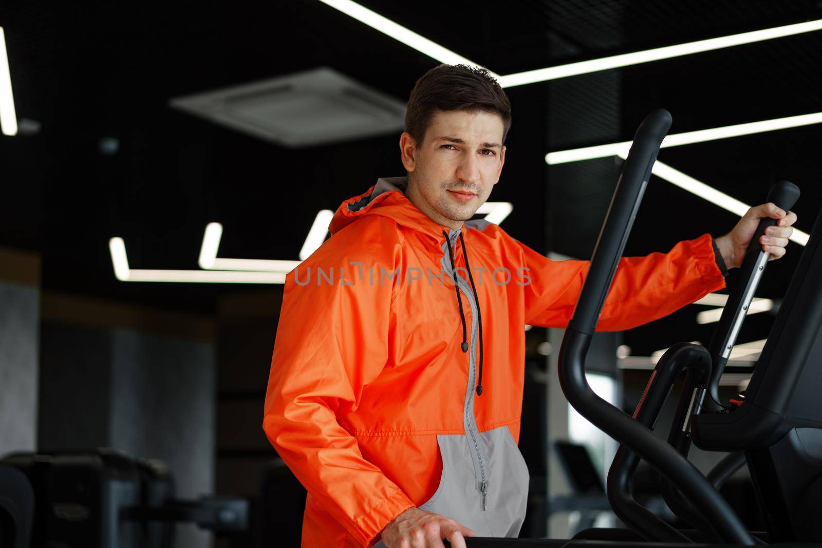 Portrait of a young man in orange windbreaker workout on a fitness machine at gym. by Fabrikasimf