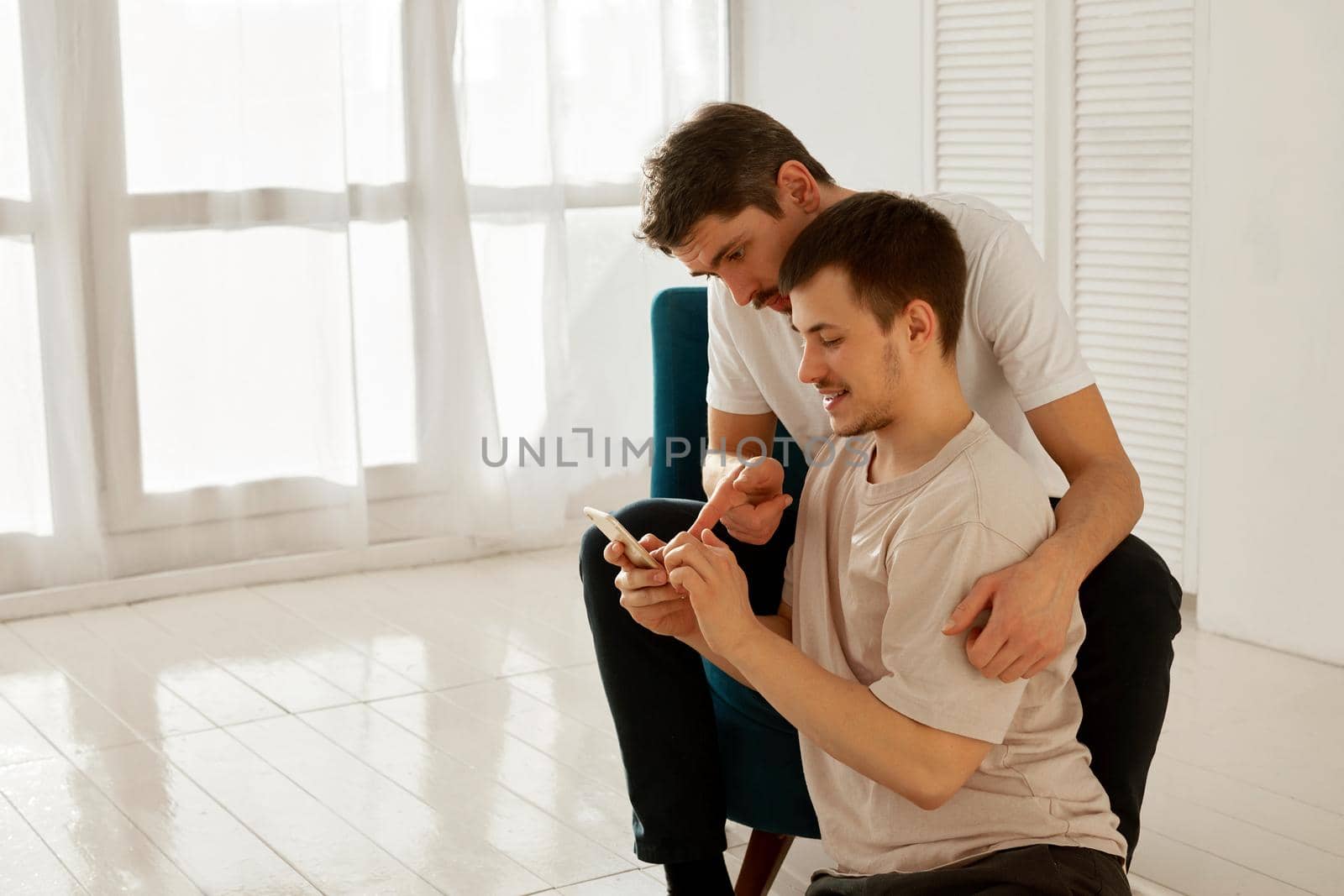 Young man hugging boyfriend and pointing at smartphone while sitting in armchair in spacious cozy room