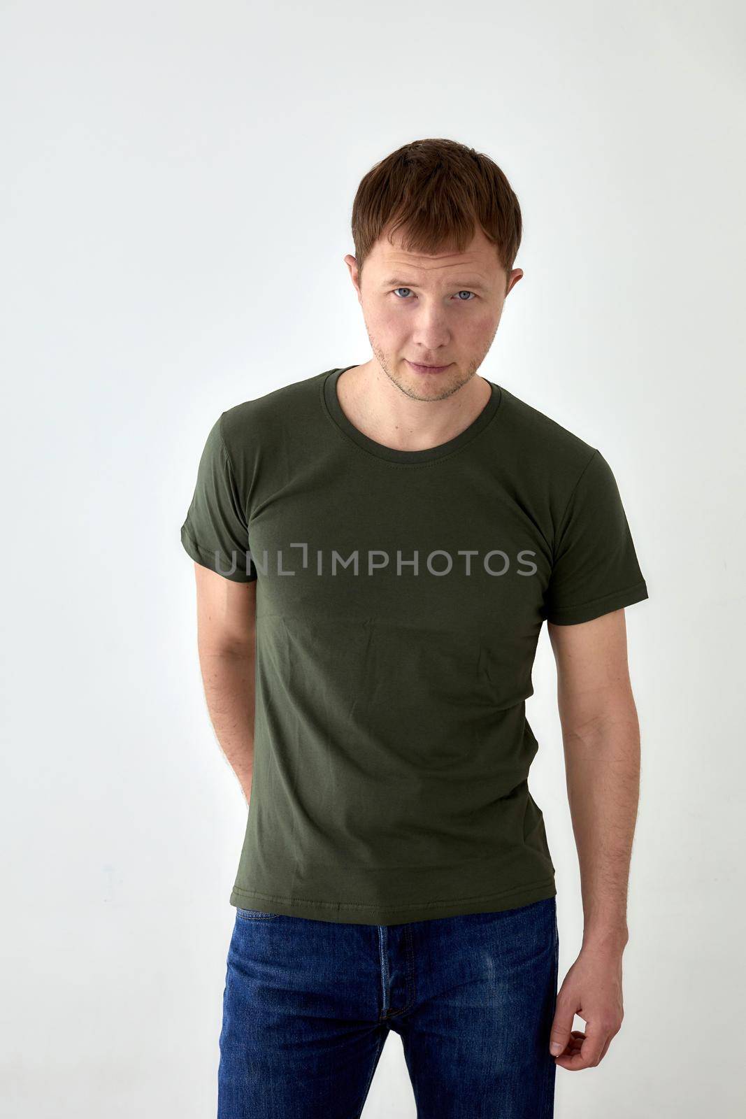 Handsome young male in jeans with hand behind and dark green t shirt standing against white background and looking at camera