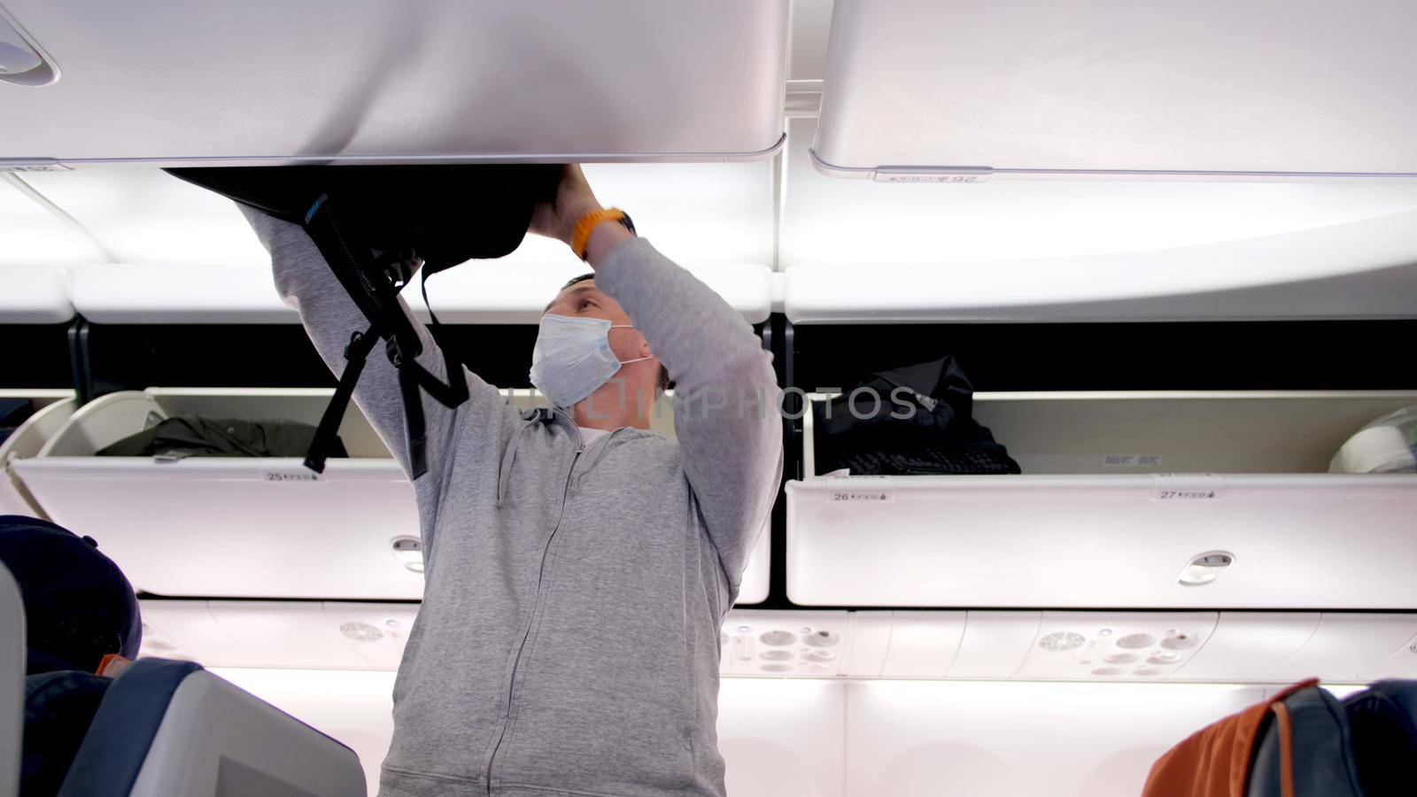 Man with protective mask in hoodie puts large black backpack on luggage rack above seats in contemporary aircraft passenger cabin