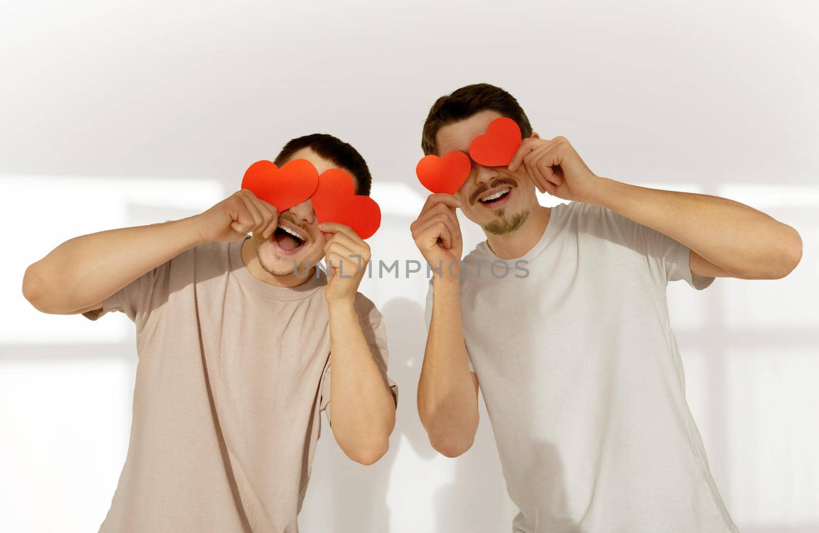 Happy couple of young gay men hide their eyes behind red paper valentine hearts in celebration at home against white wall background
