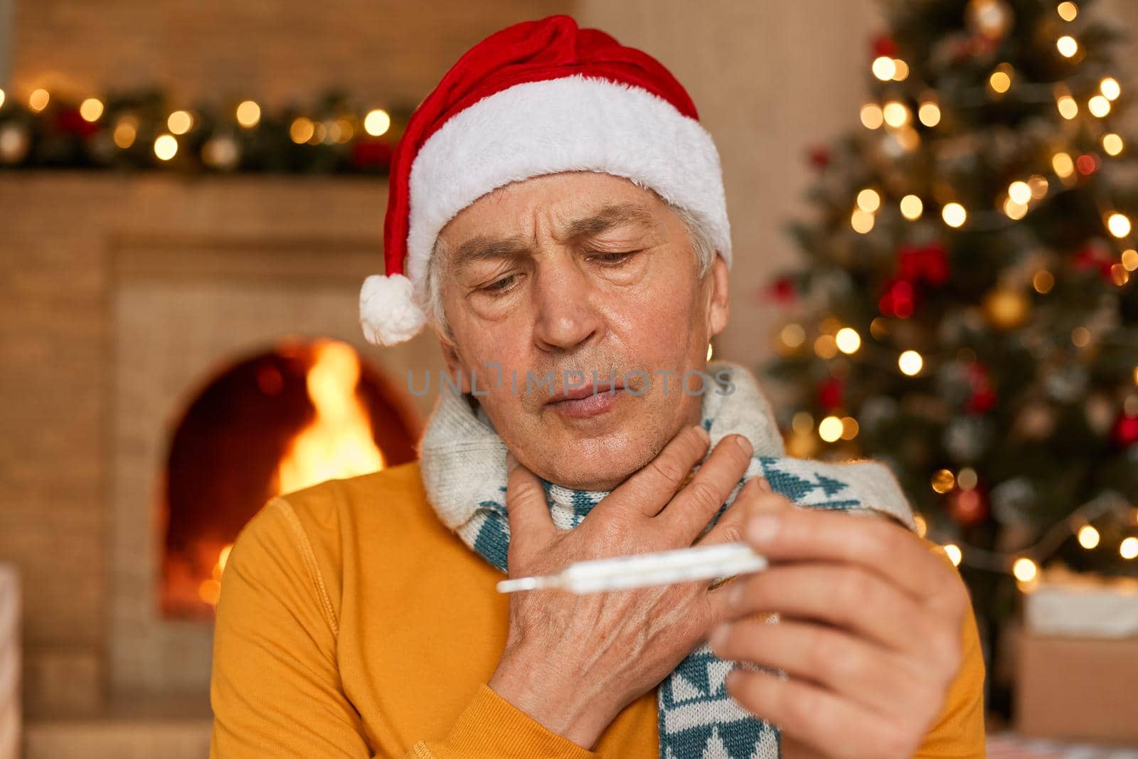 Sick senior man in santa hat sitting near fireplace wrapped in scarf, wearing santa claus hat, holding thermometer in hands, has fever, suffers from sore throat, poses room with Christmas decoration.
