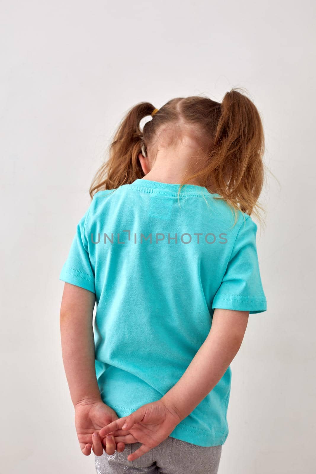 Back view of unrecognizable upset little girl with ponytails dressed in blue t shirt standing against white wall