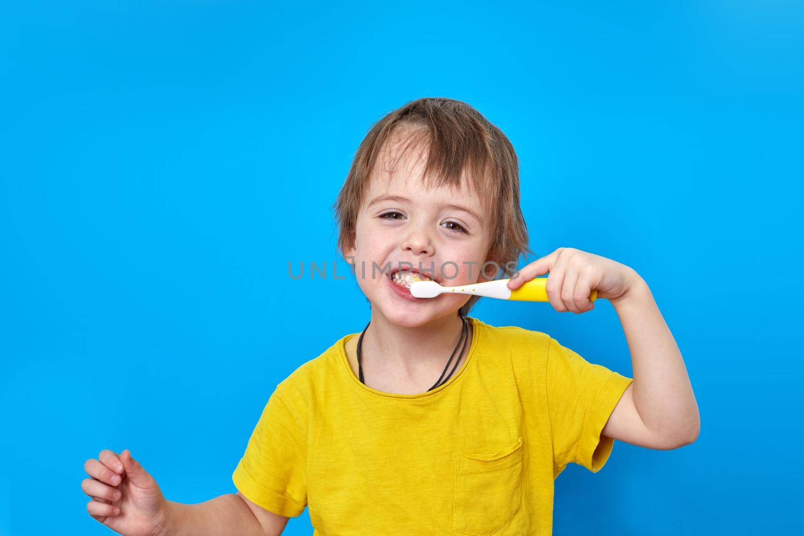 Smiling handsome little boy is brushing his teeth with brush on blue background